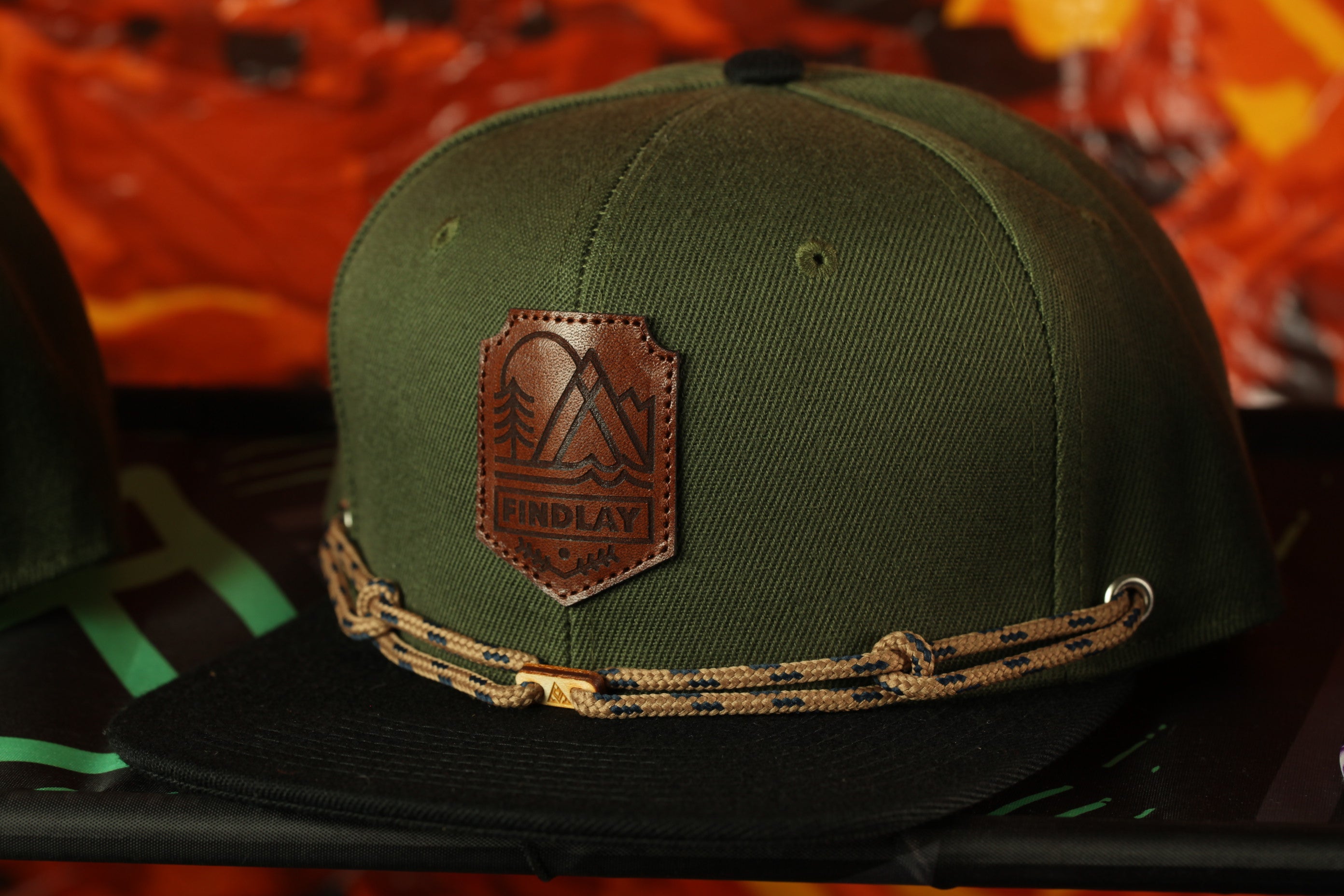 A green baseball cap with a leather patch and a braided strap, placed on a black surface against a colorful background.