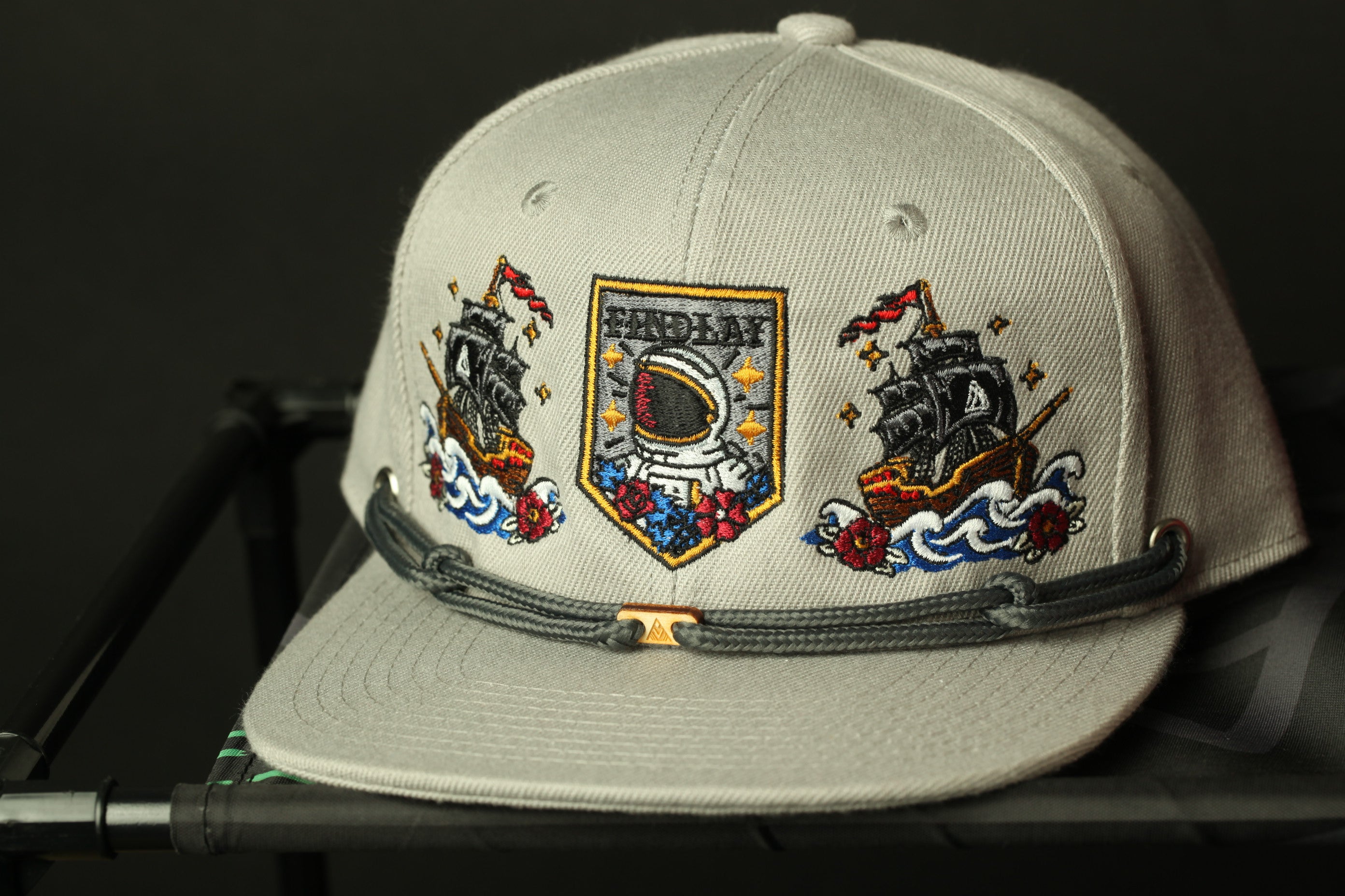 A gray baseball cap with various embroidered designs, including a ship, a person in a spacesuit, and other maritime-themed imagery.