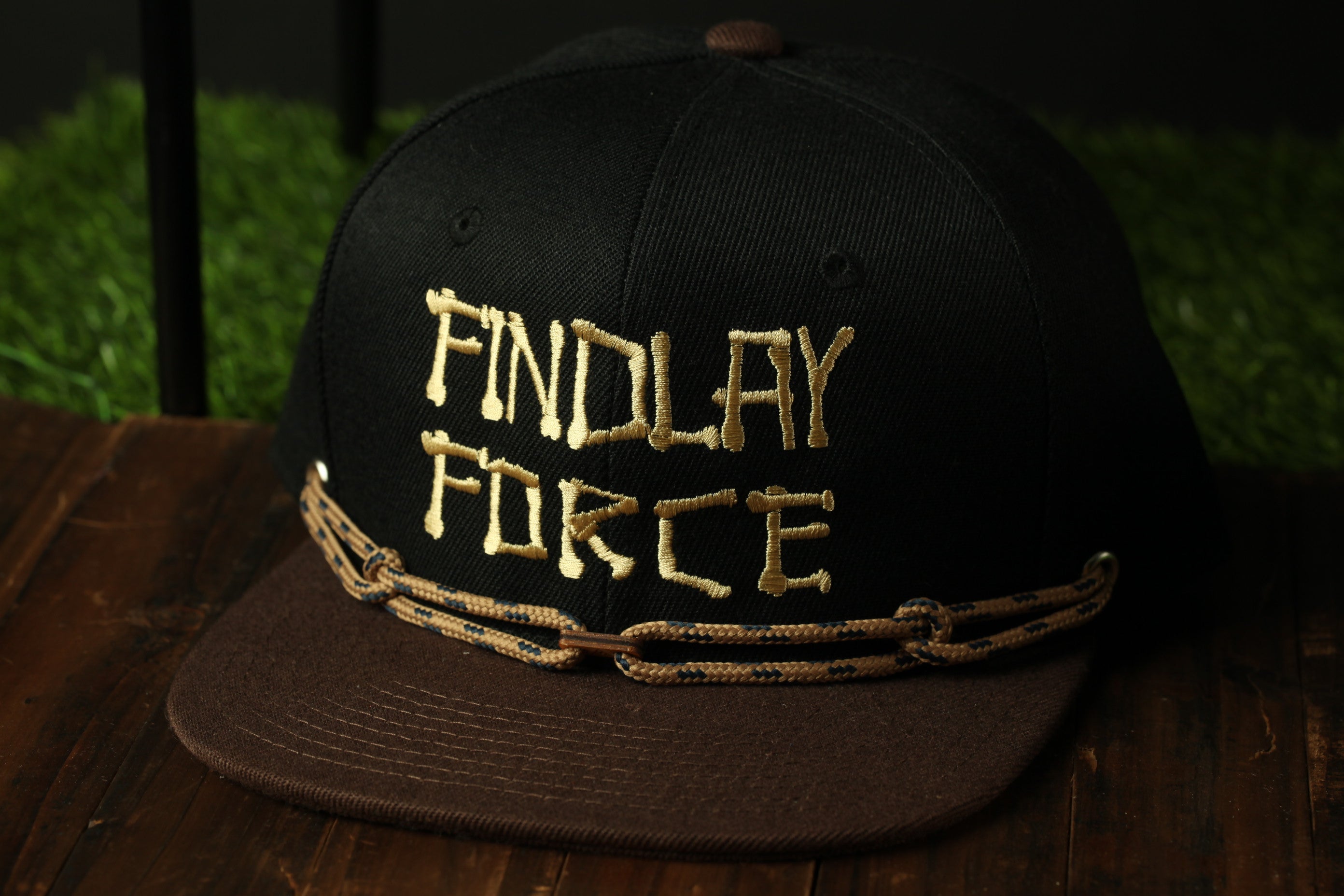 A black baseball cap with gold embroidered text on it, placed on a wooden surface.