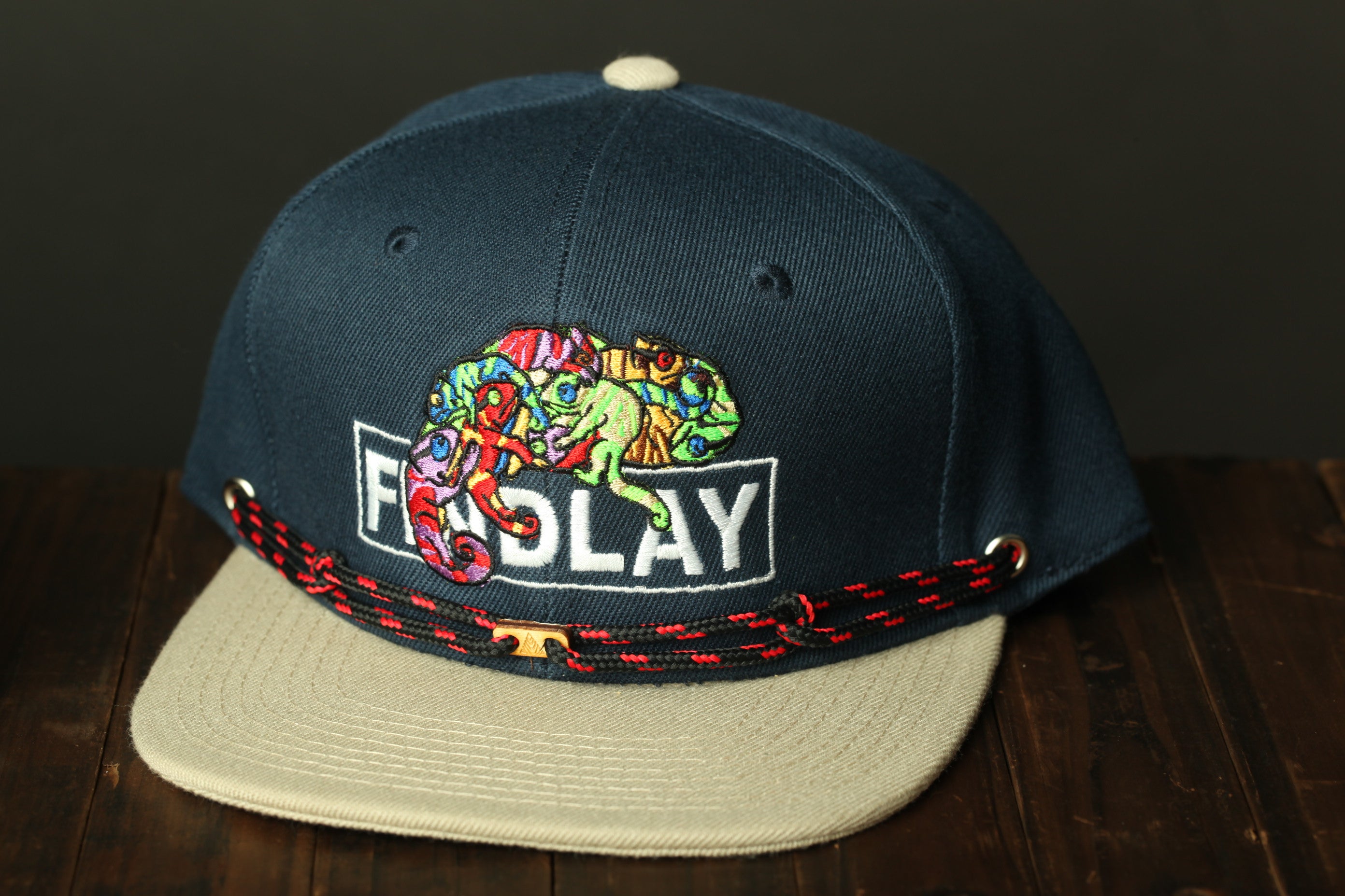 A navy blue baseball cap with a colorful chameleon design and the word "FINDlay" printed on it, sitting on a wooden surface.