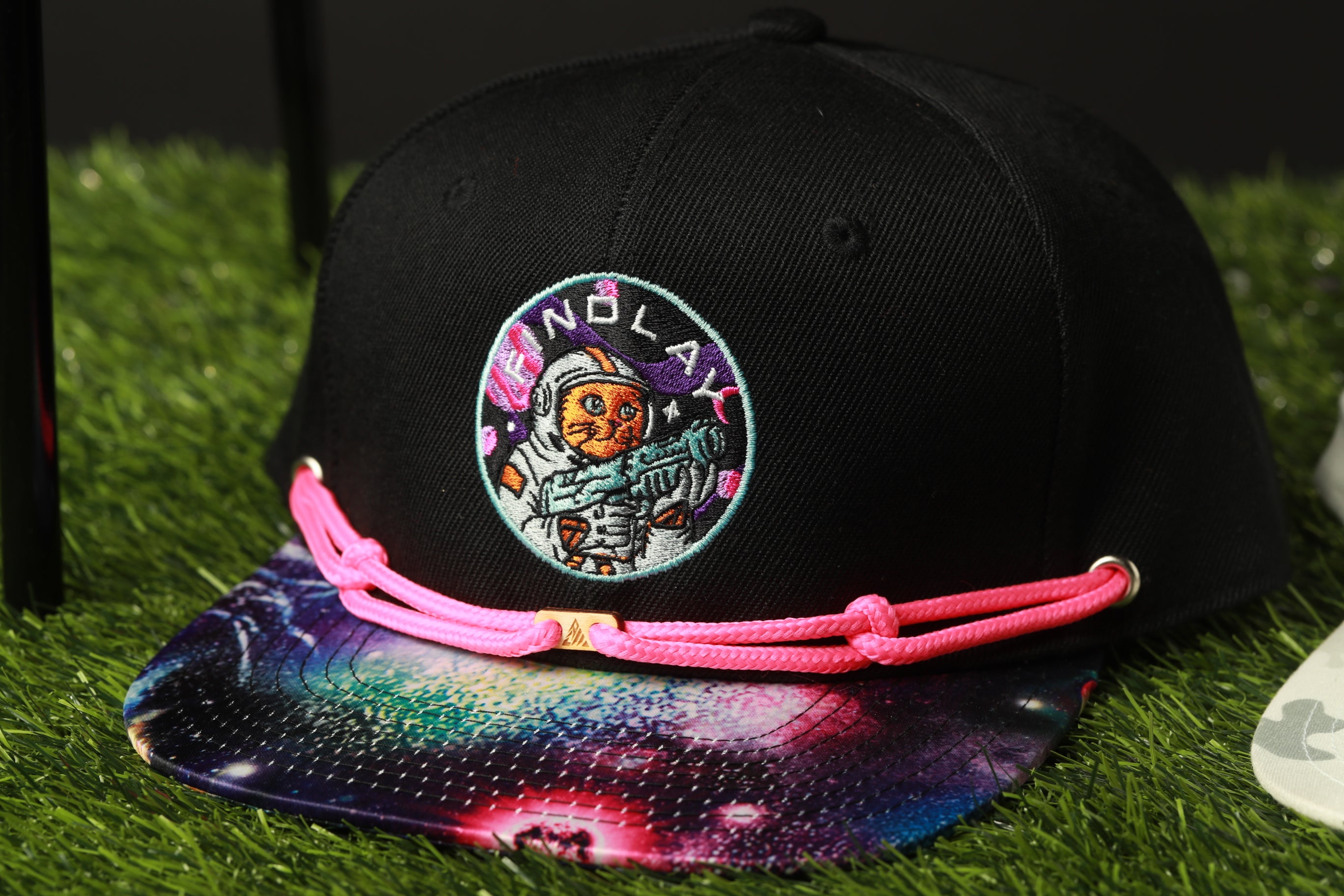 A black baseball cap with a colorful, space-themed design and a pink braided strap.