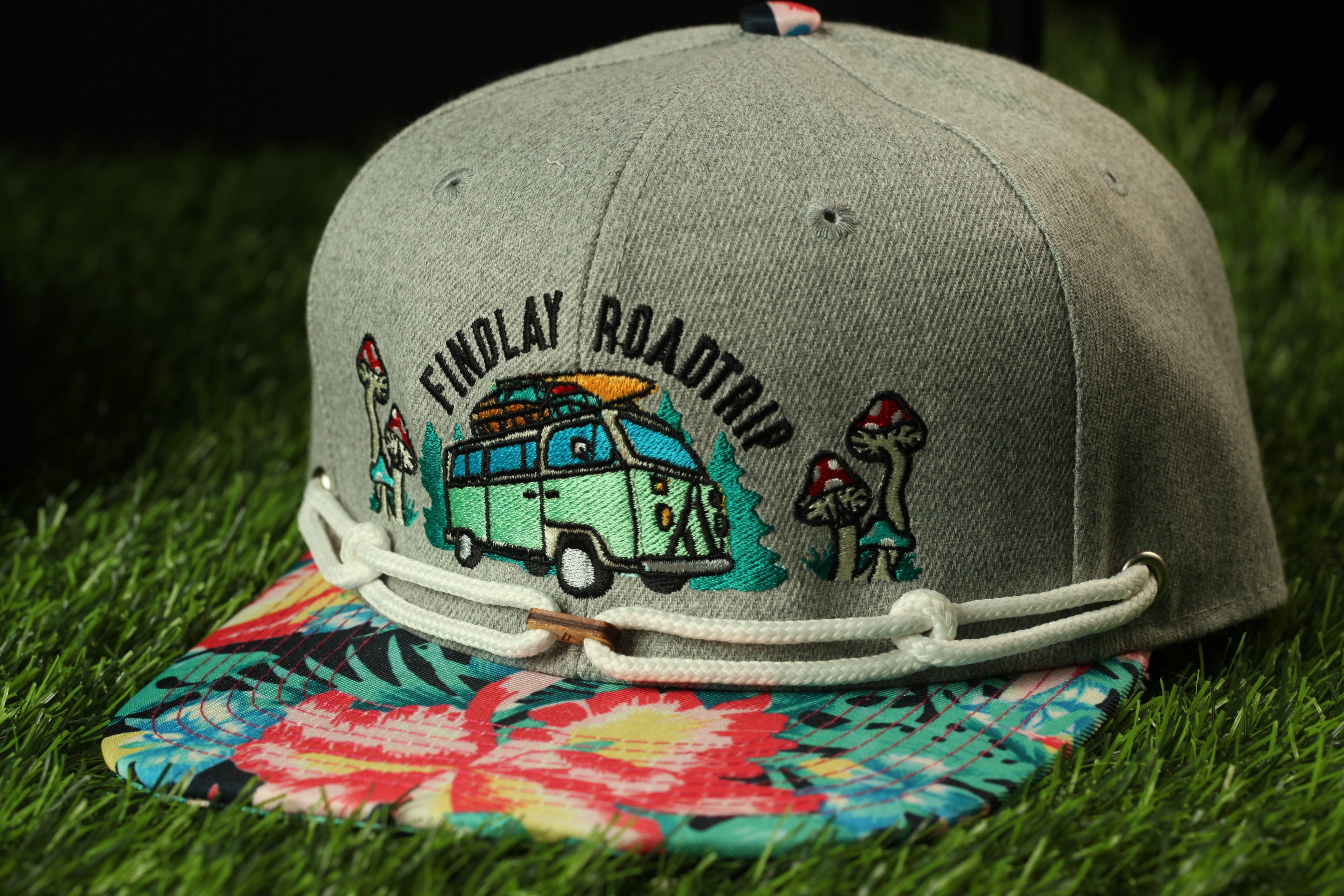 A gray baseball cap with a colorful floral pattern and a cartoon van design, sitting on a grassy surface.