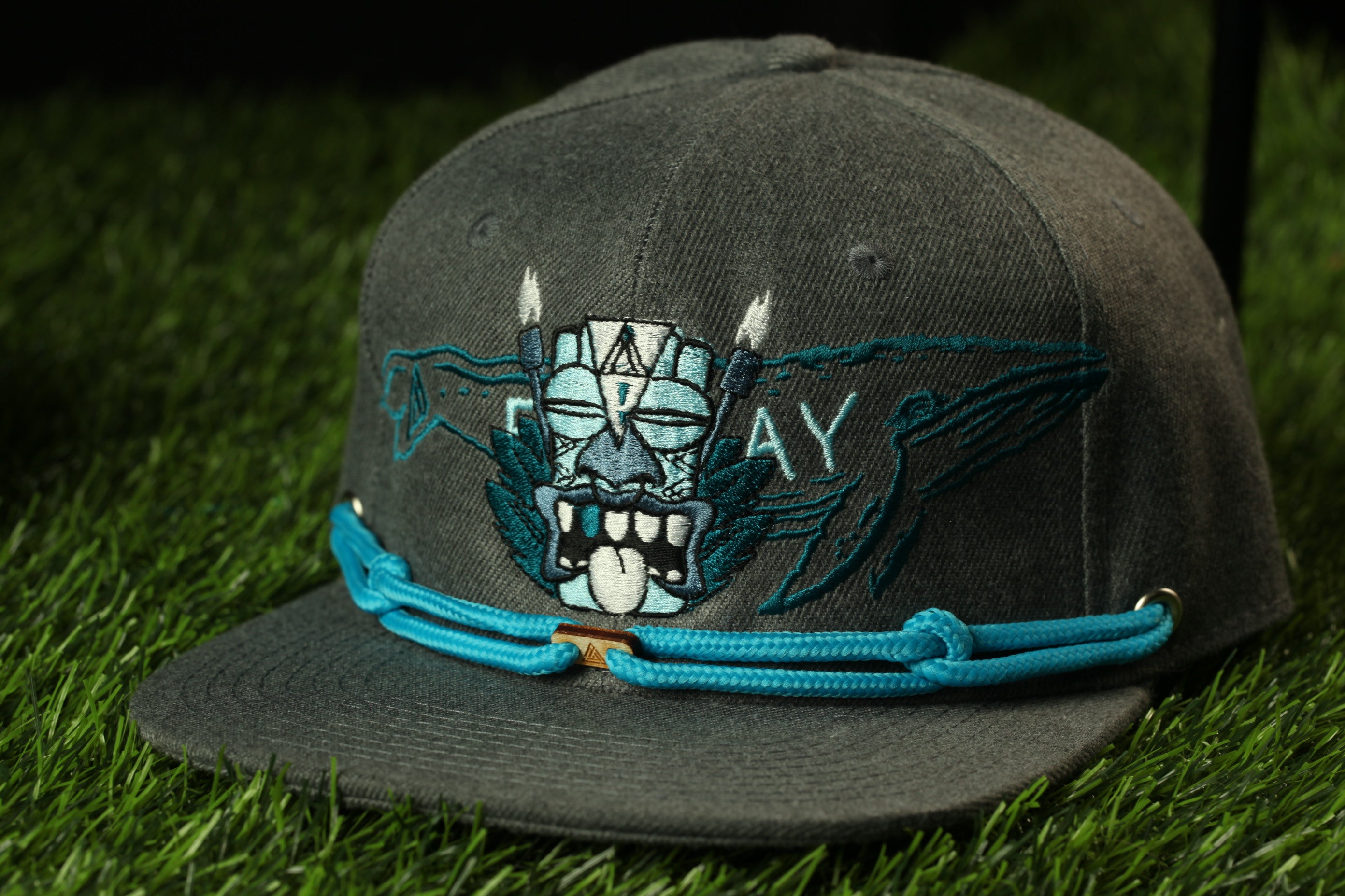 A gray baseball cap with a blue string and a graphic design on the front, sitting on a grassy surface.