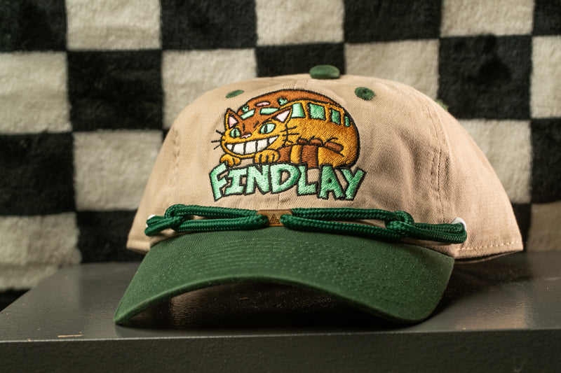 A beige baseball cap with a green brim and a cat embroidered on the front, sitting on a black surface against a checkered background.