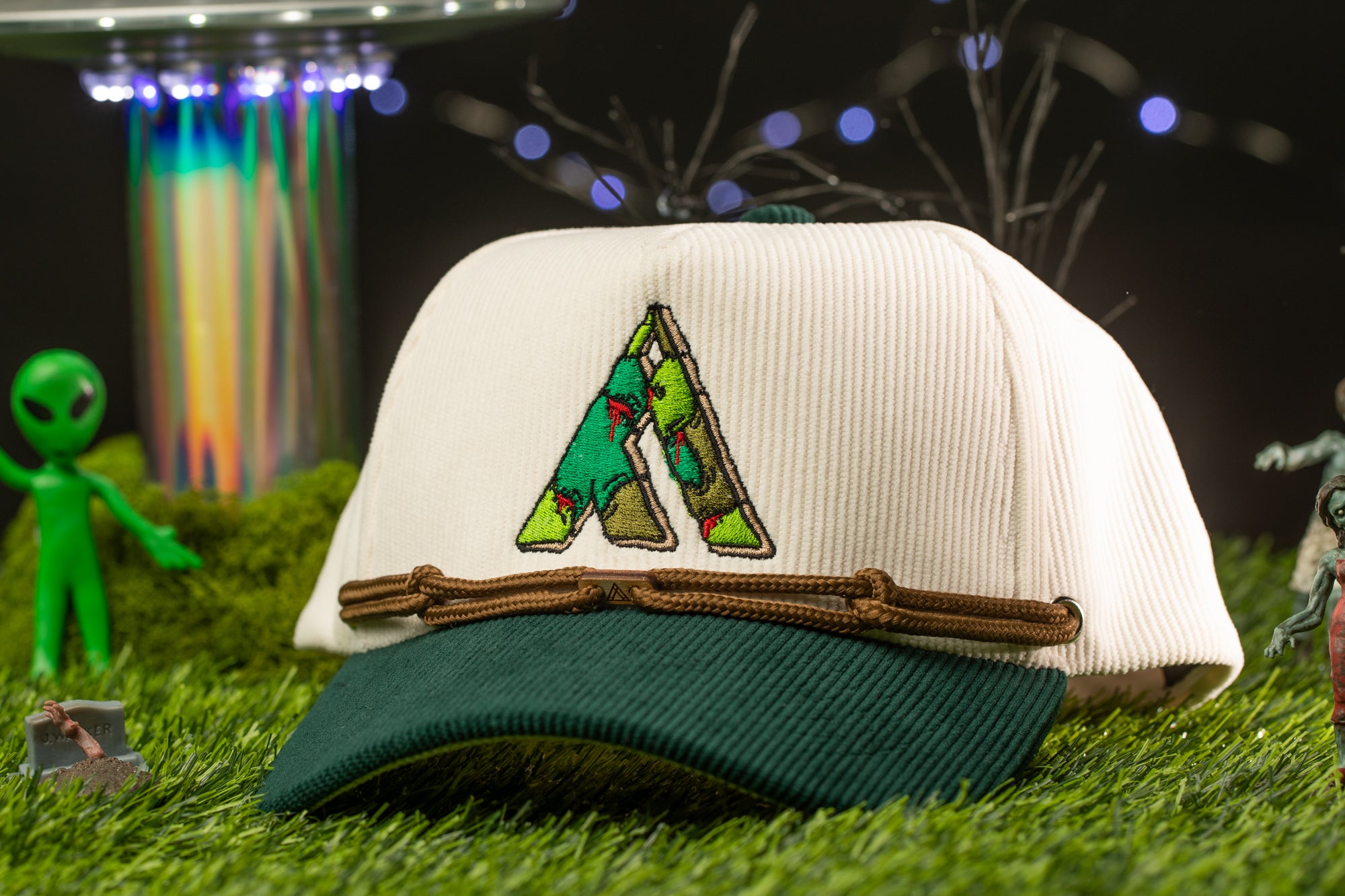 A white baseball cap with a green brim and a green alien figure in the background, set against a grassy surface with a colorful, illuminated structure in the background.