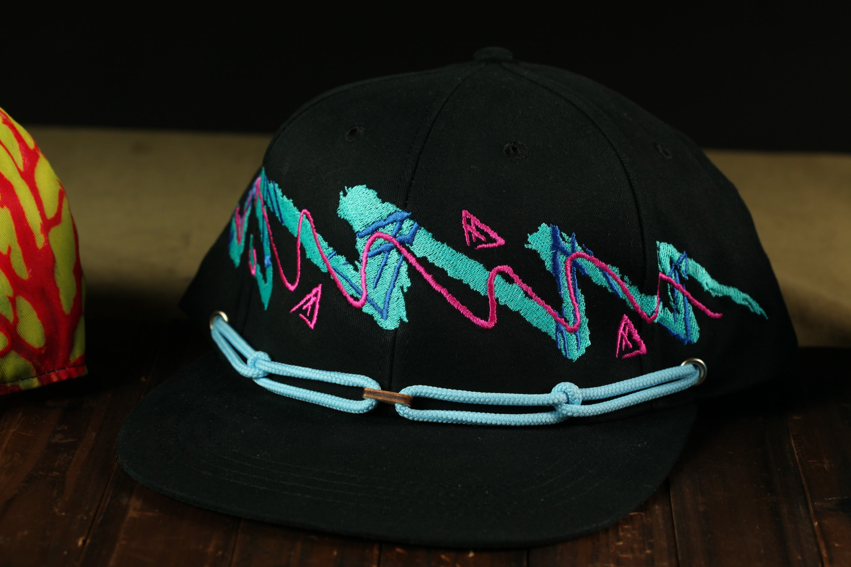 A black baseball cap with colorful geometric patterns and a blue chain strap is displayed on a wooden surface.