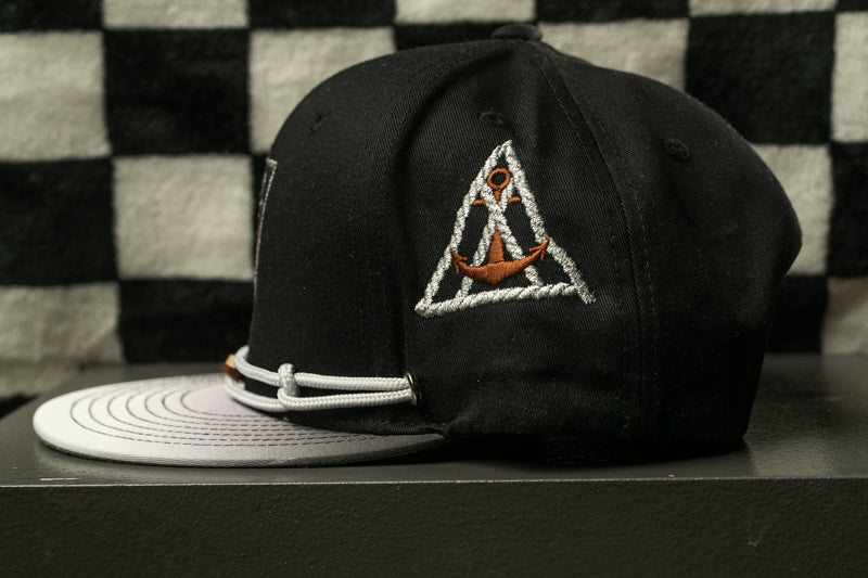A black baseball cap with a white brim and a white circular band, featuring an embroidered triangle with an anchor symbol on the side.