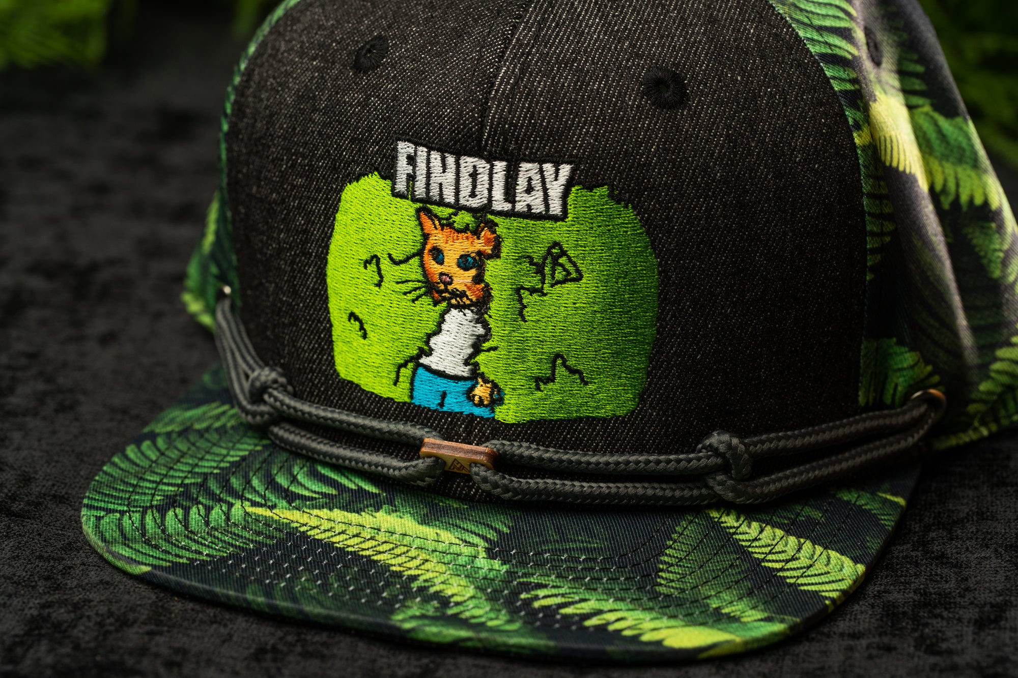 A black baseball cap with a green and black camouflage pattern, featuring a cartoon image of a cat and the word "FINDLAY" printed on it.