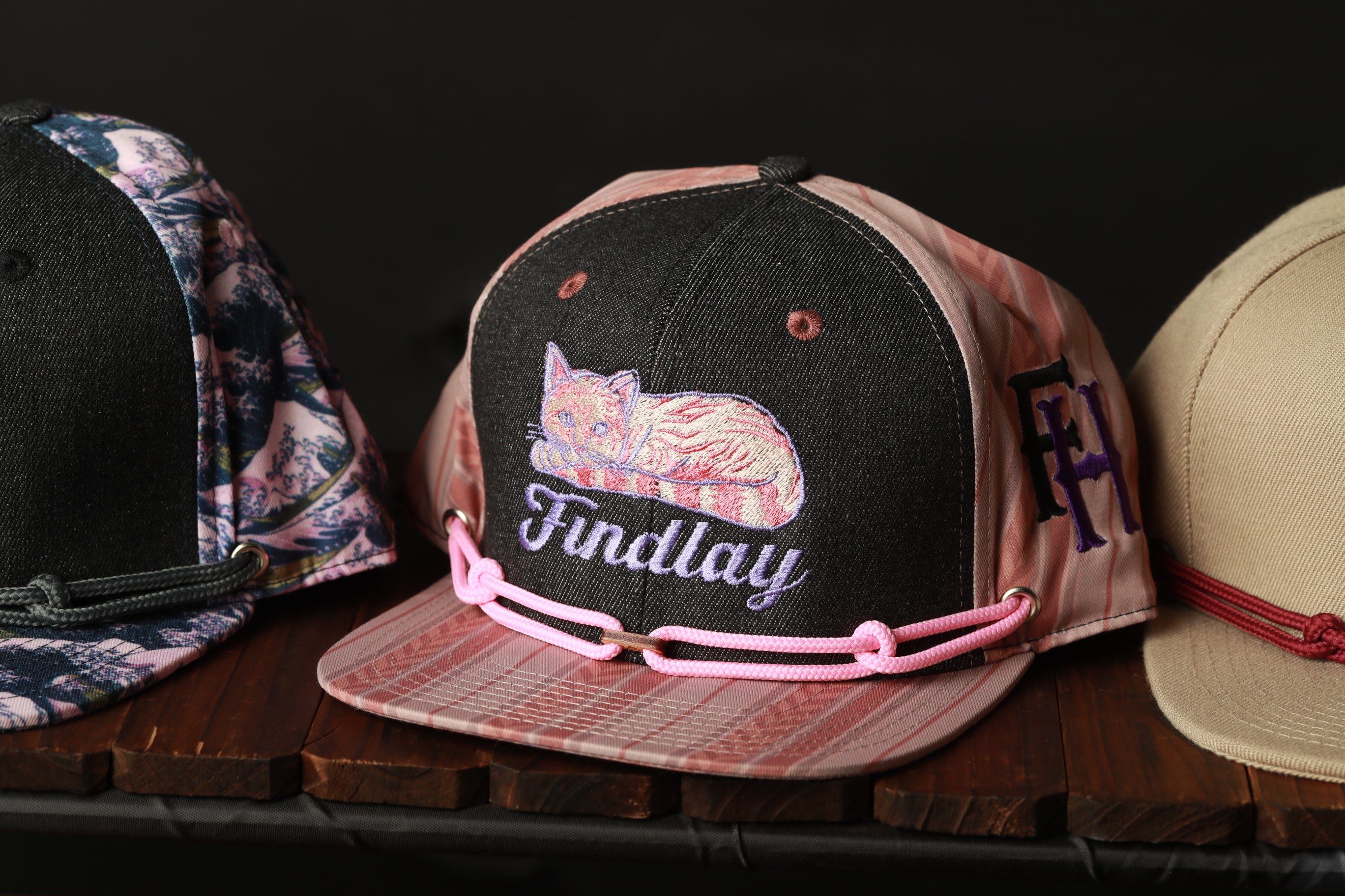 Three baseball caps are displayed on a wooden surface, with one featuring a cat design and the other two having different patterns.