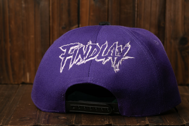 A purple baseball cap with the word "FINDA" printed on it, sitting on a wooden surface.
