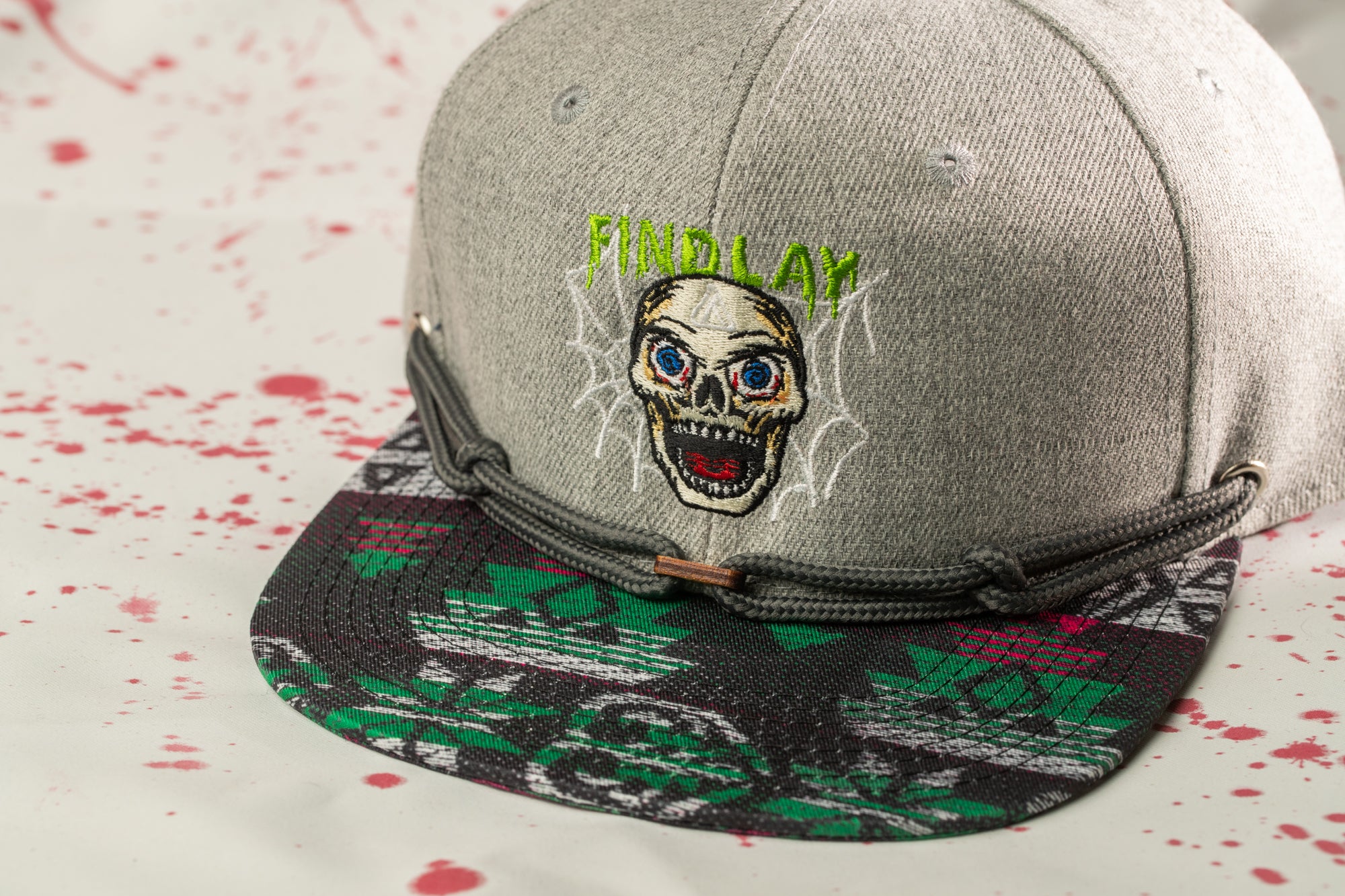 A gray baseball cap with a skull design and the text "FINELY LAUGH" printed on it, placed on a surface with red splatters.