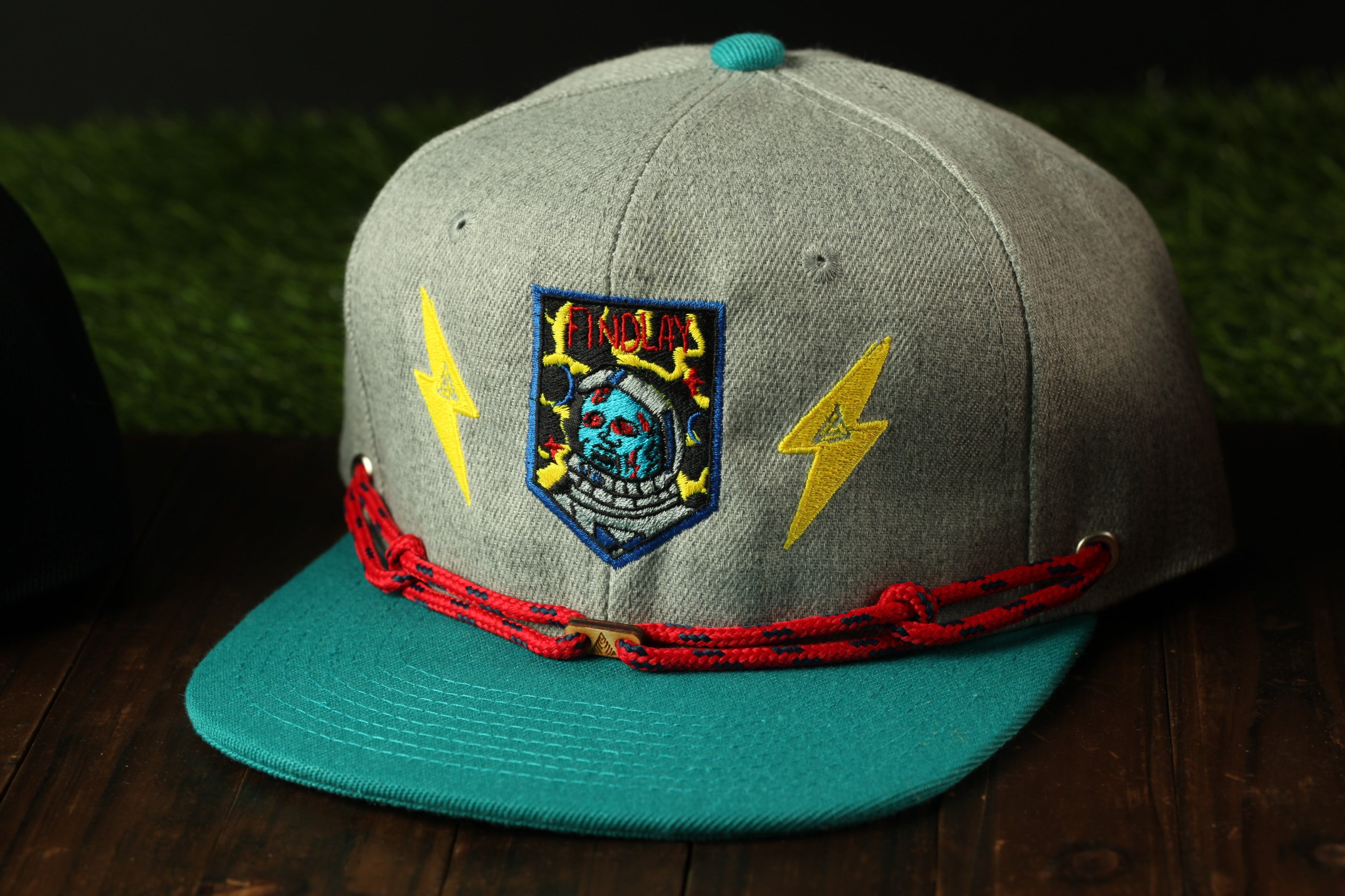 A gray baseball cap with a teal brim and red braided strap, featuring a patch with a skull and lightning bolt design.