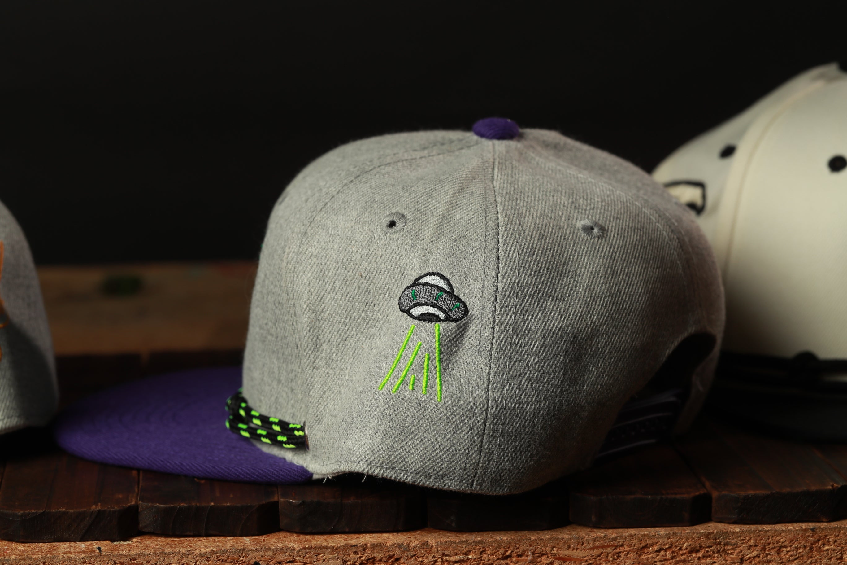 A gray baseball cap with a purple brim and a green alien embroidered on the front, sitting on a wooden surface.