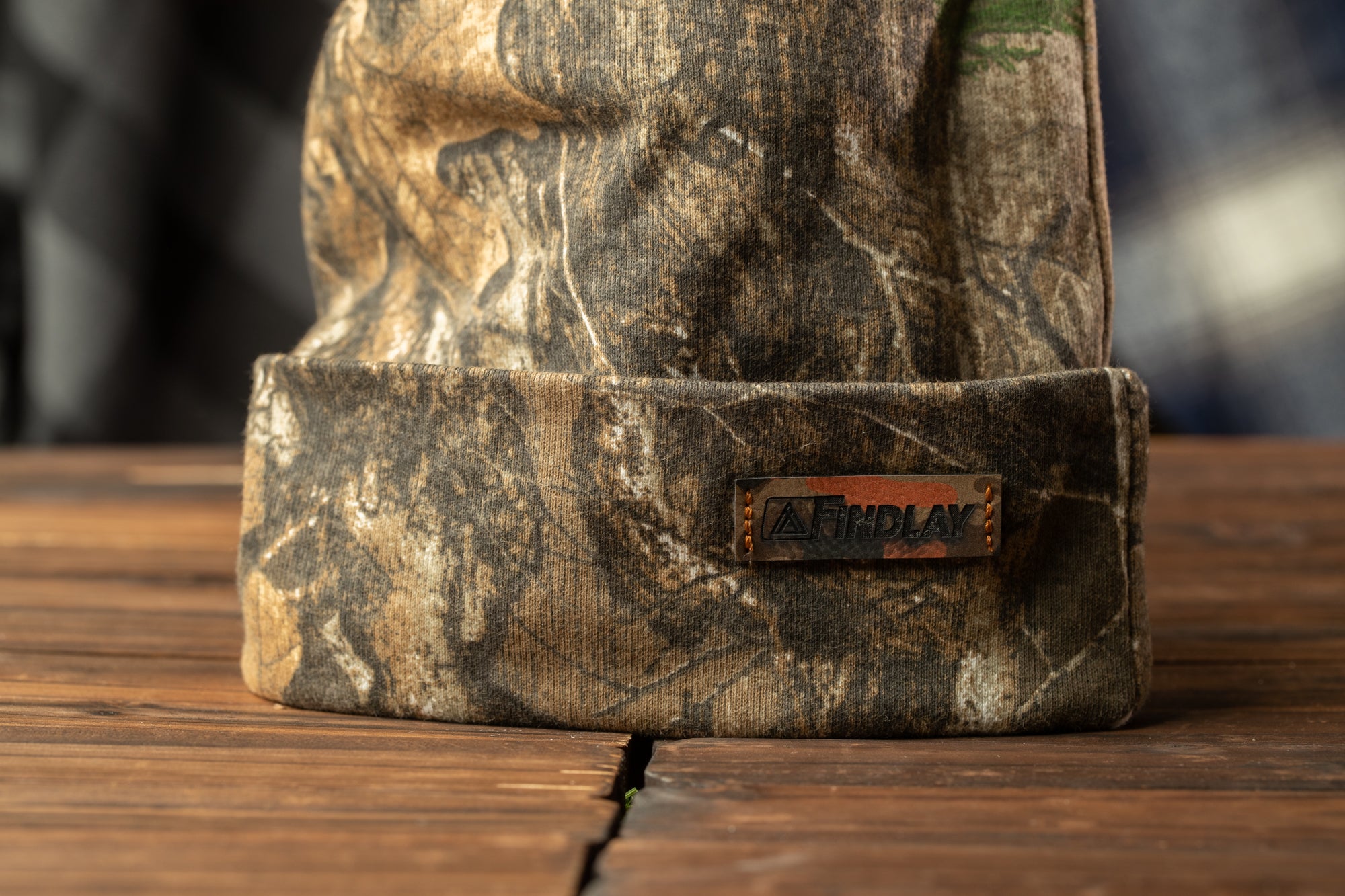 A camouflage-patterned beanie with a metal "FINDLAY" label attached to it, placed on a wooden surface.
