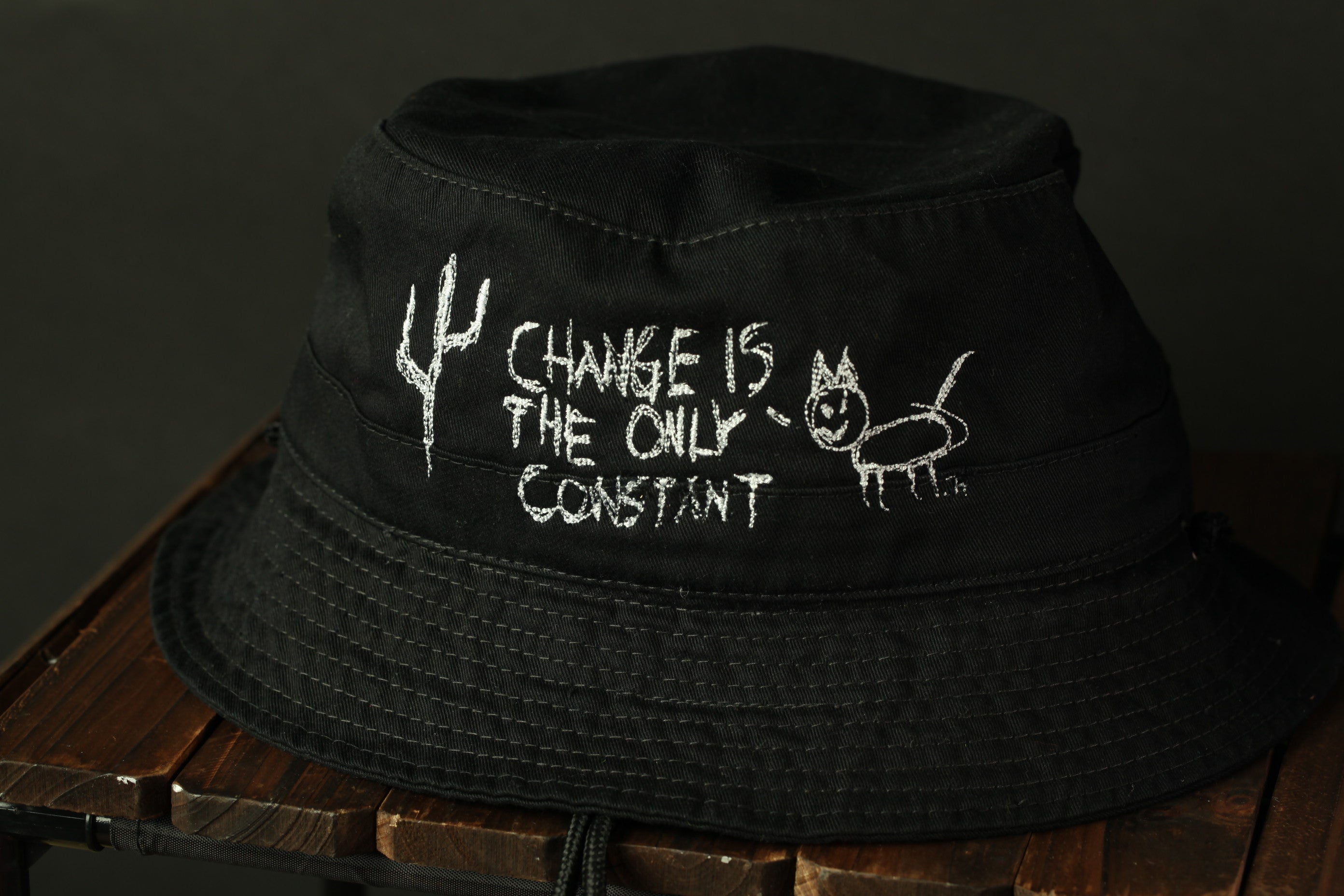 A black bucket hat with white text and a drawing on it, placed on a wooden surface.