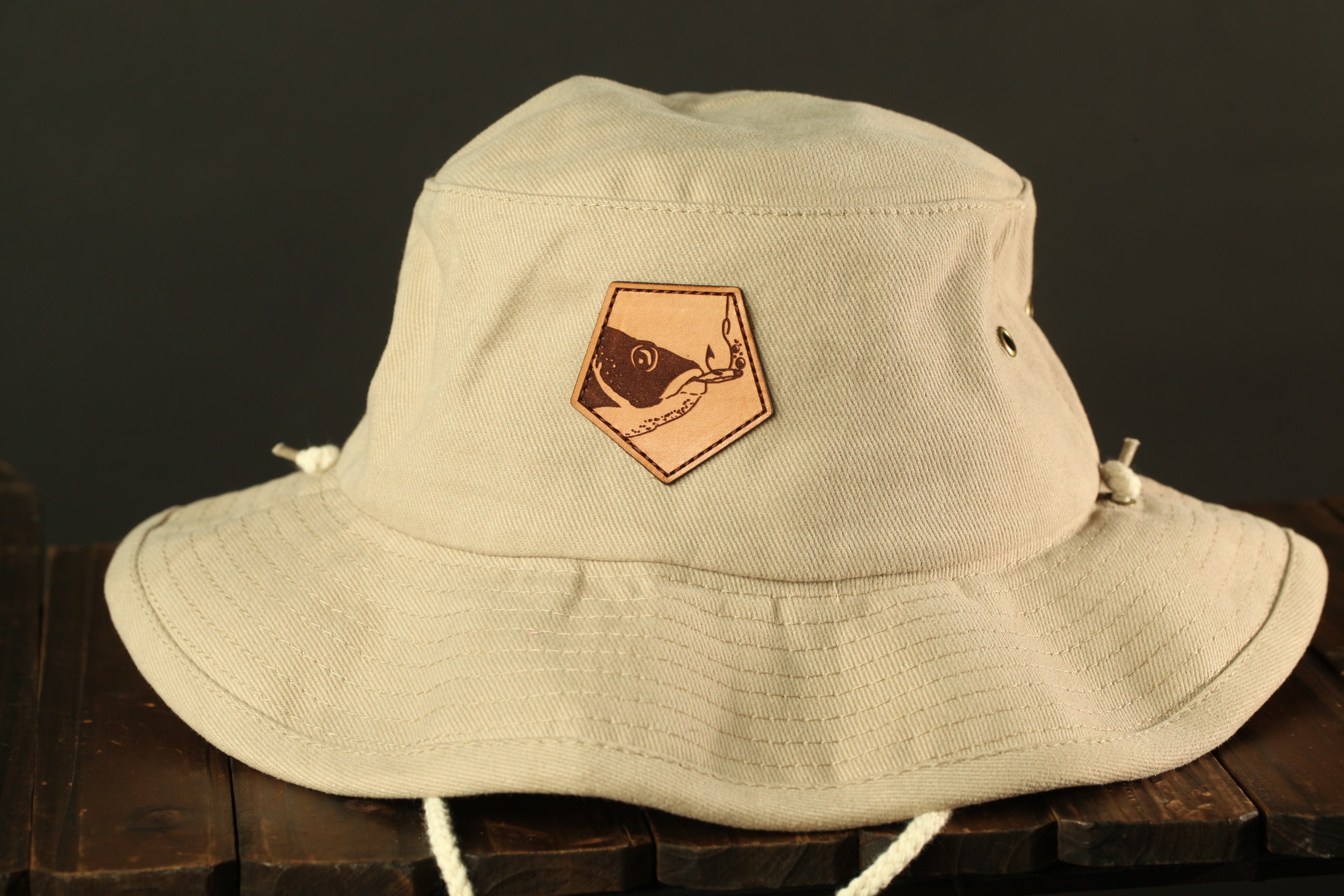 A beige bucket hat with a leather patch on the front, sitting on a wooden surface.