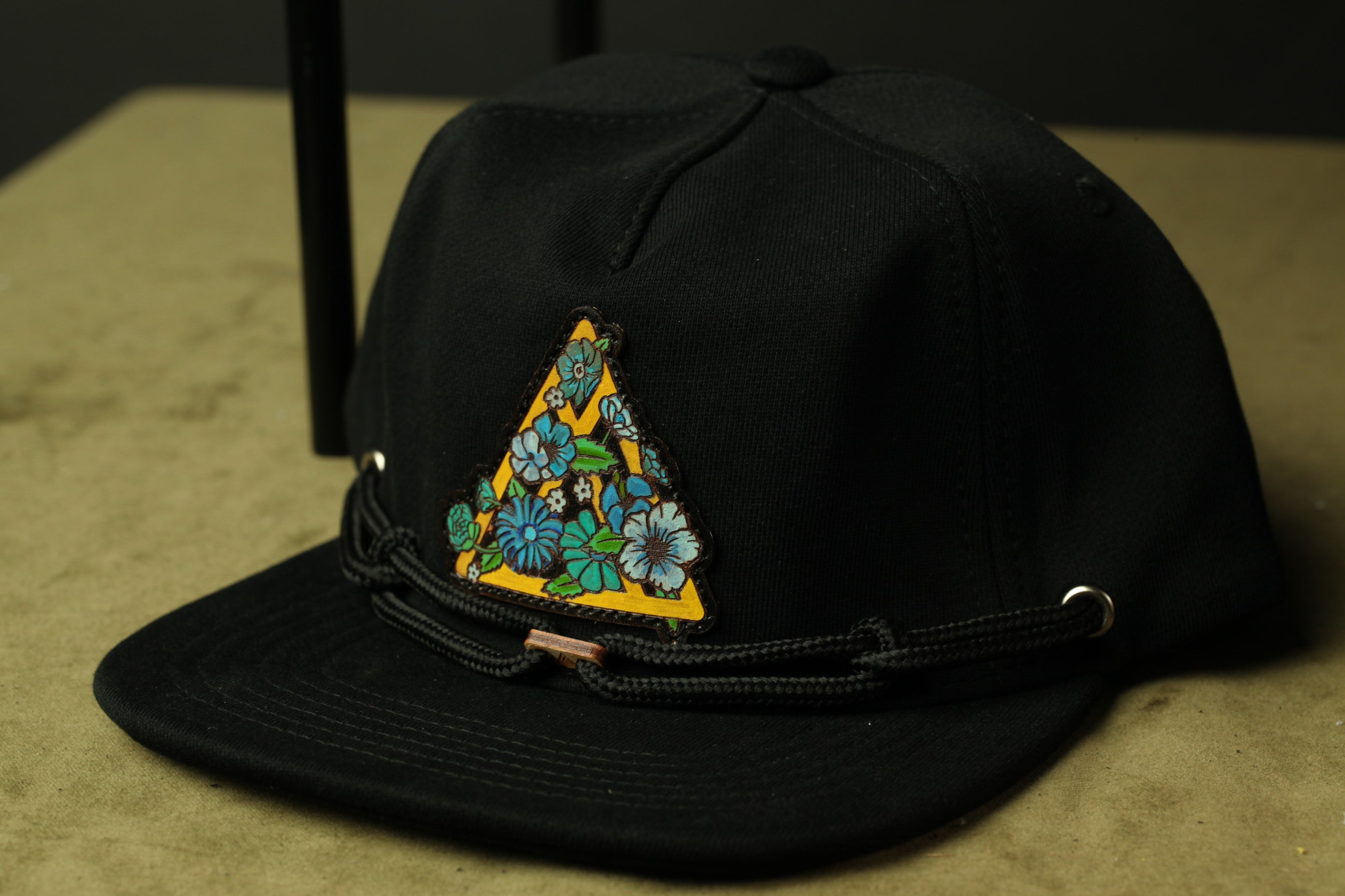 A black baseball cap with a yellow triangle design featuring blue and green flowers on the front.