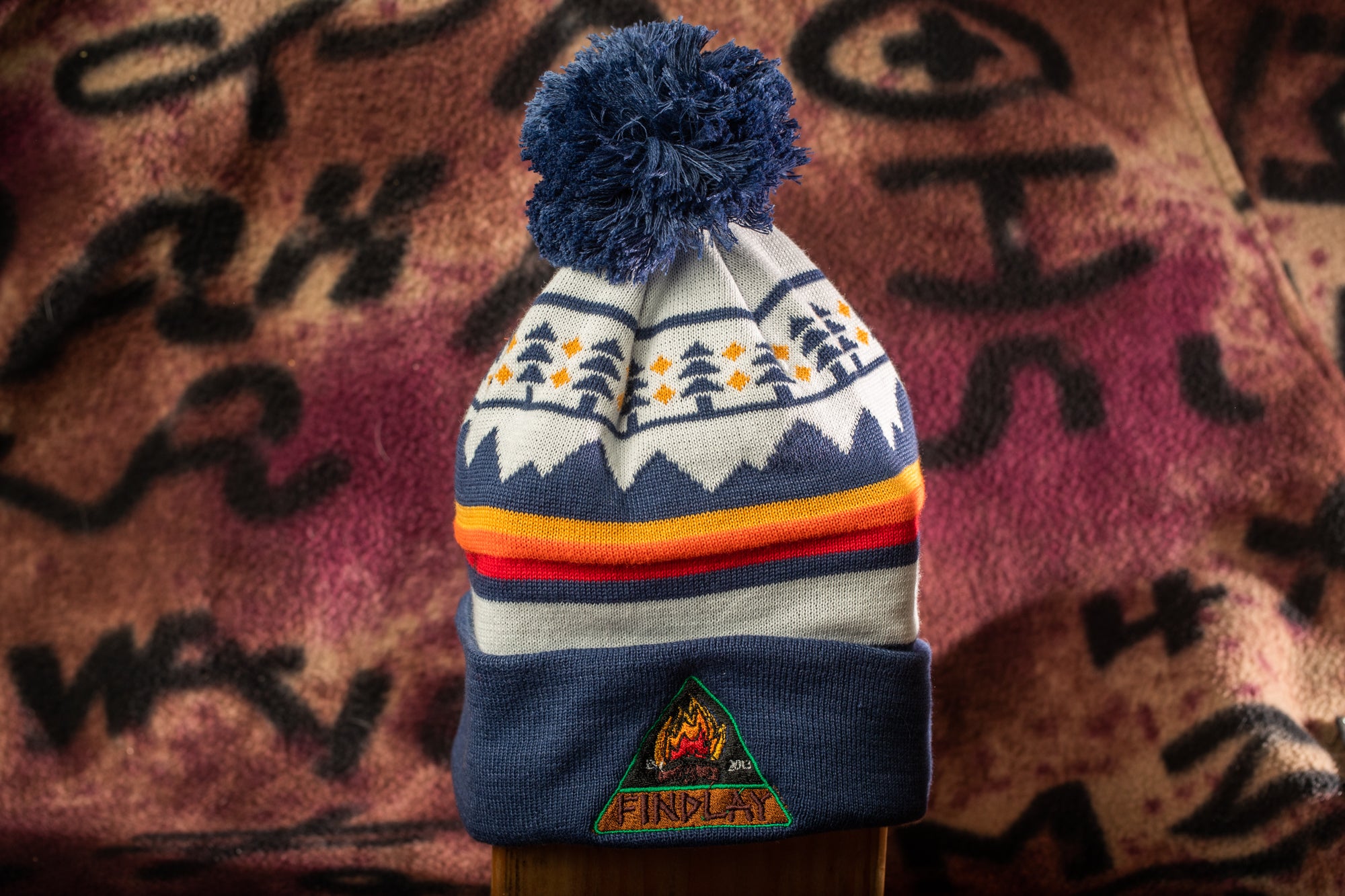 A colorful winter hat with a pom pom and a logo, placed against a patterned background.
