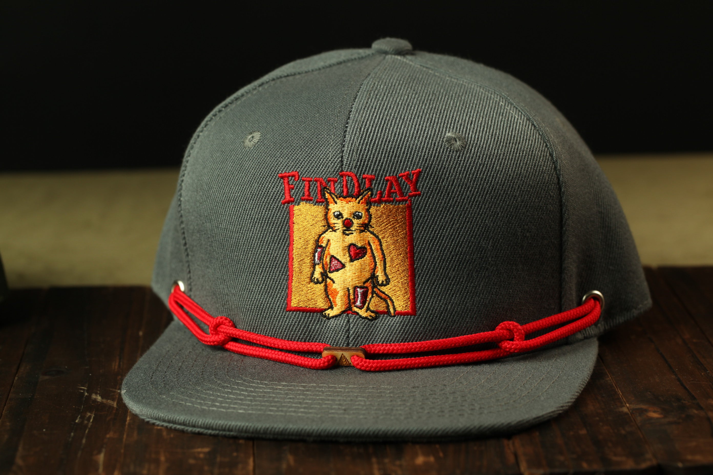 A gray baseball cap with a red string and a cat embroidered on the front, sitting on a wooden surface.