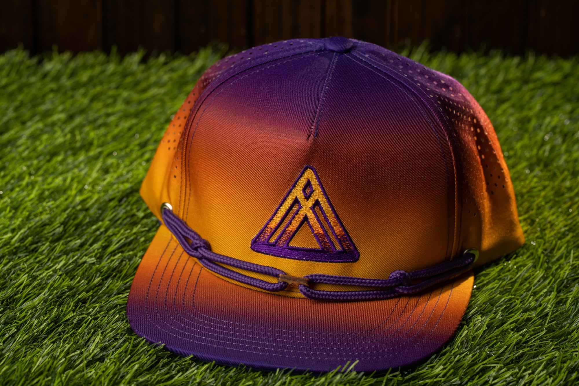A purple and orange gradient baseball cap with a triangular logo on the front, resting on a grassy surface.