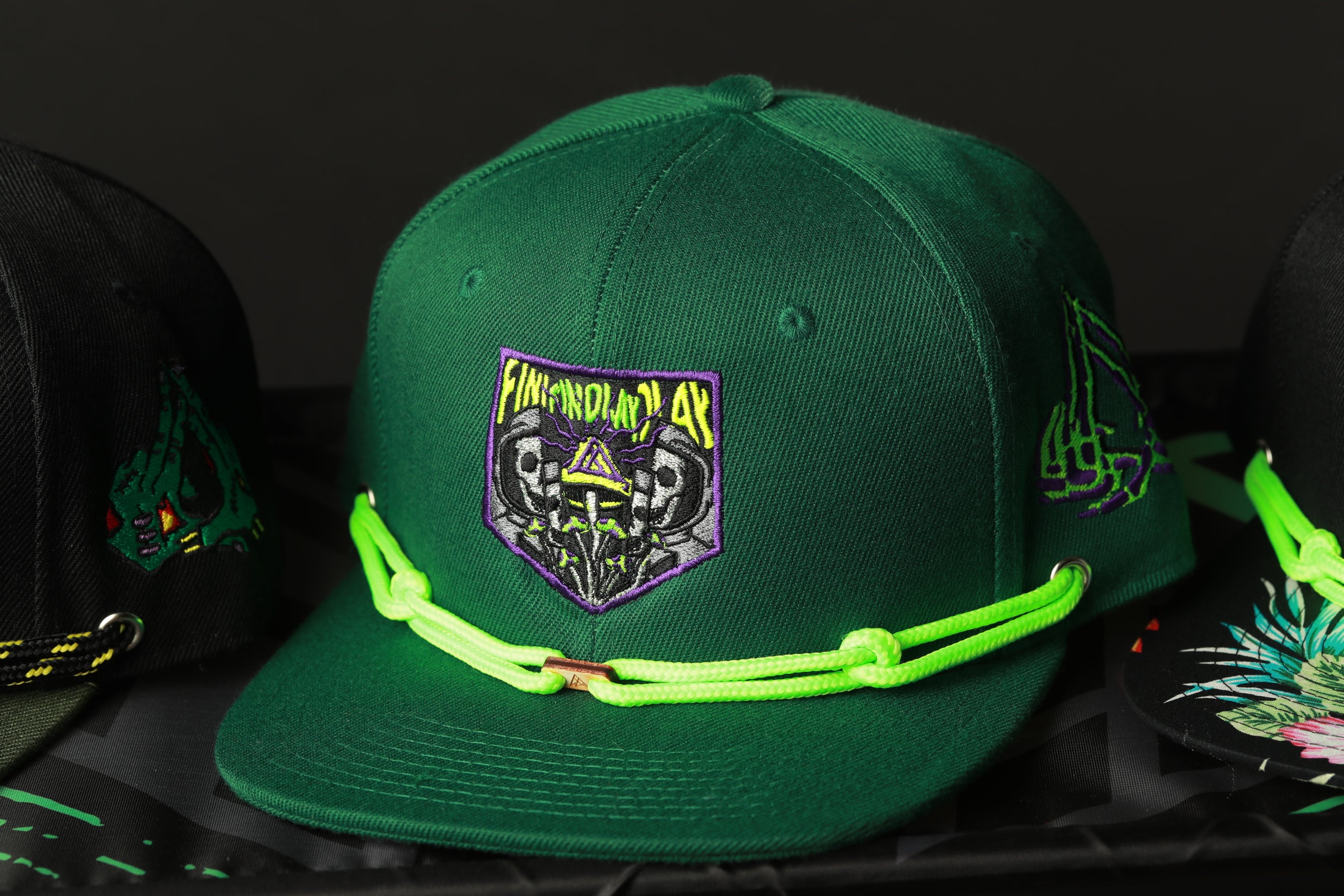 A close-up image of a green baseball cap with a purple and yellow design, featuring a skull and crossbones motif, and neon green strings.