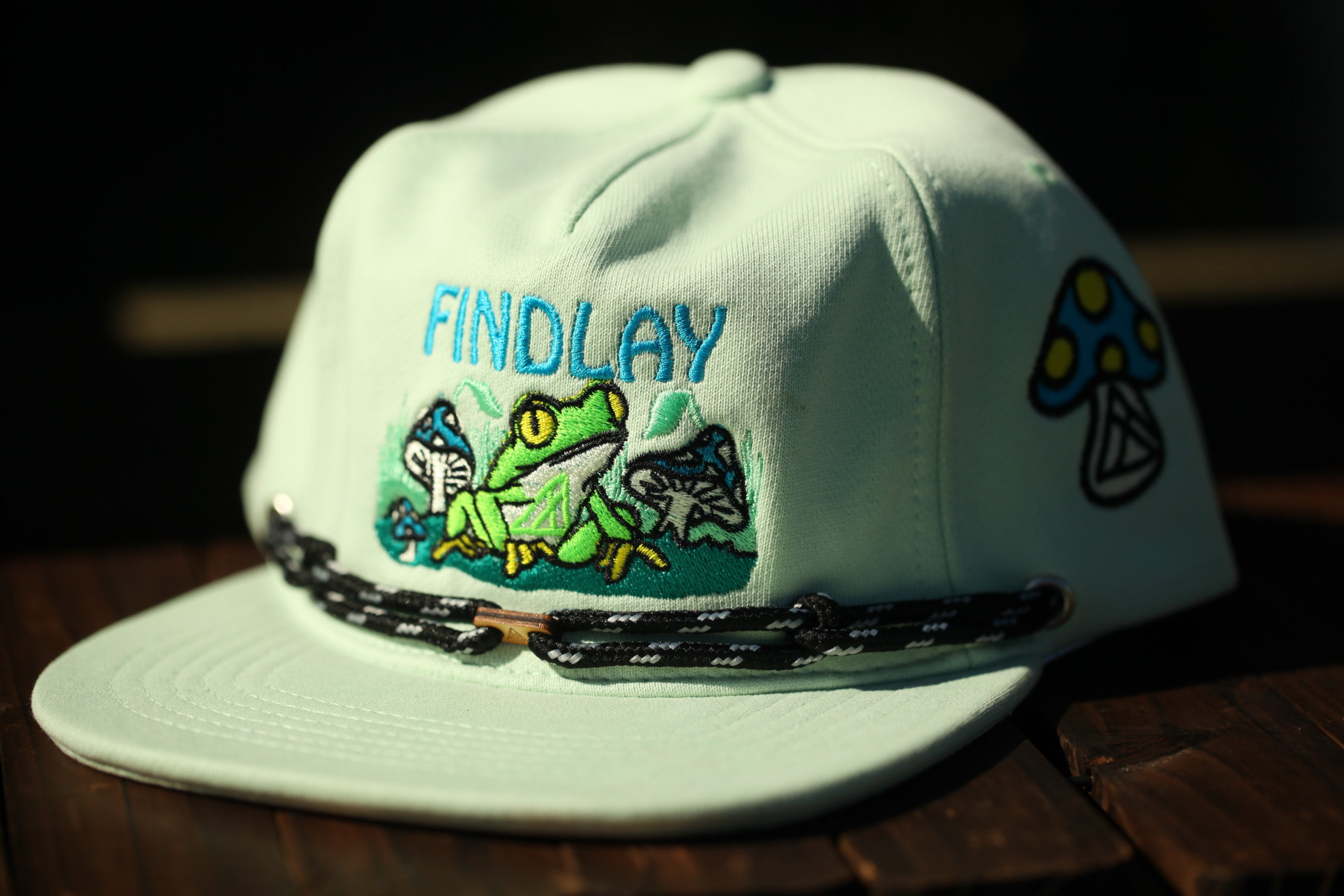 A white baseball cap with a green frog and mushroom design, featuring the word "FINDLAY" on the front.