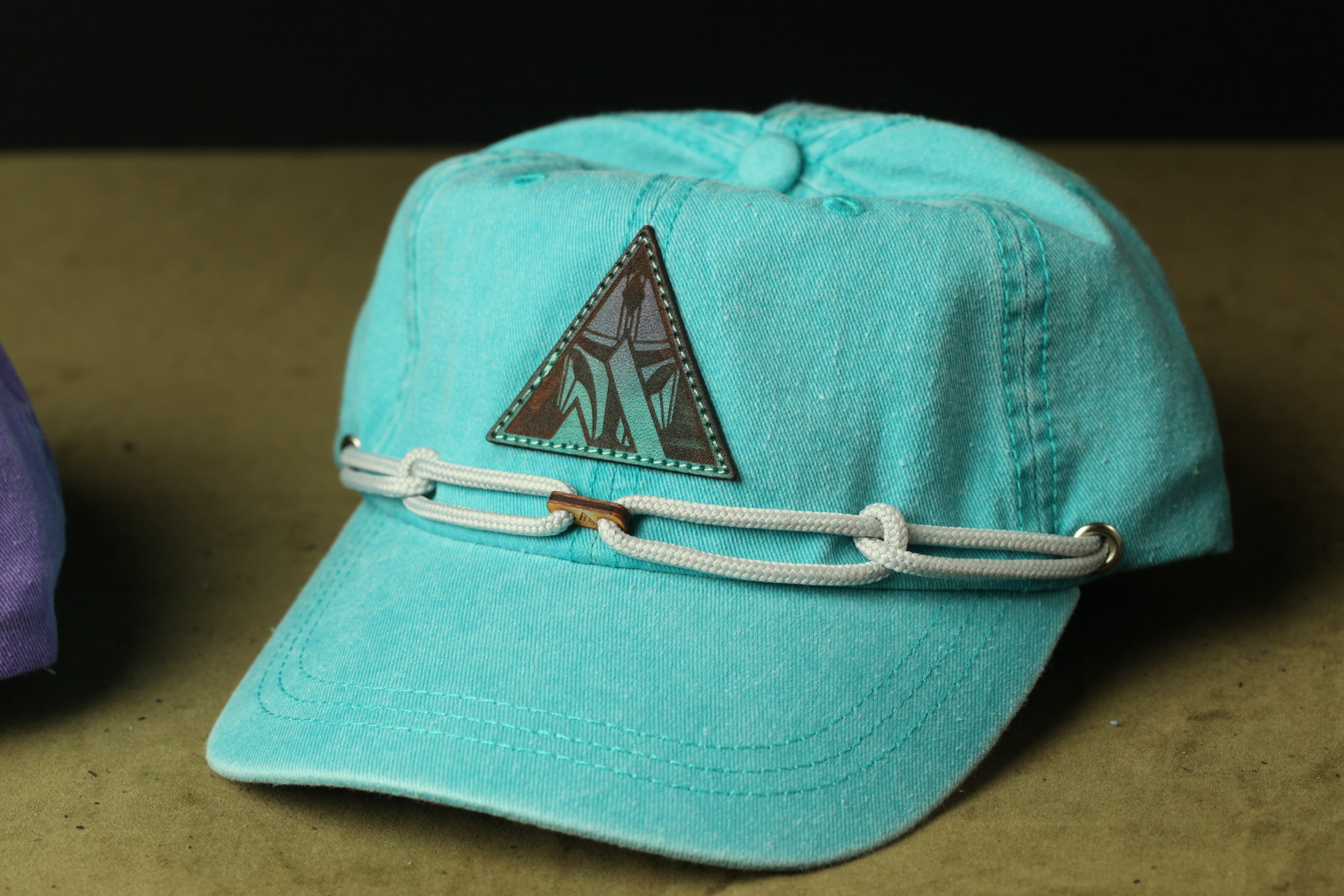 A turquoise baseball cap with a triangular patch on the front and a white rope or chain strap.