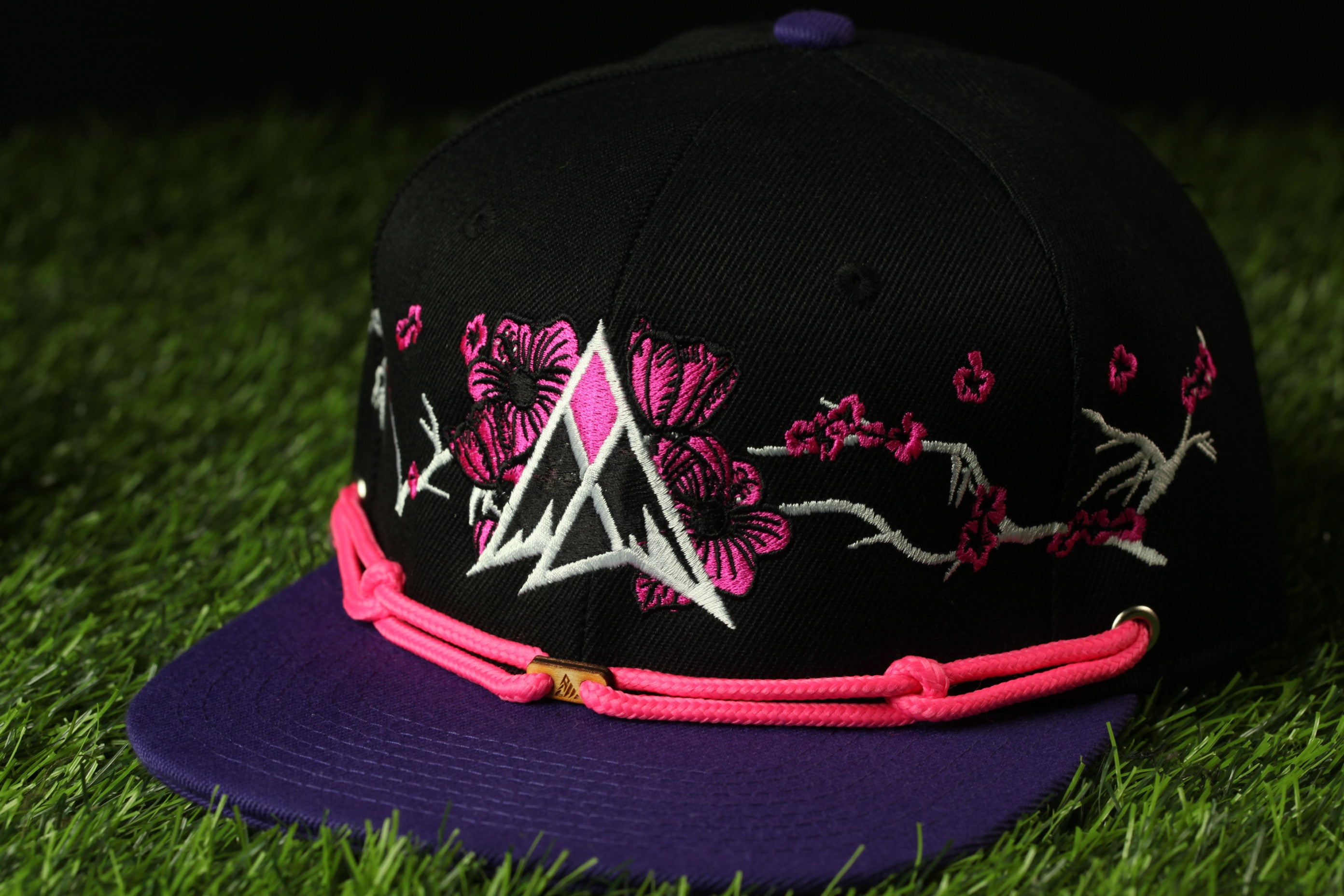 A black baseball cap with pink floral embroidery and a purple brim, resting on a grassy surface.