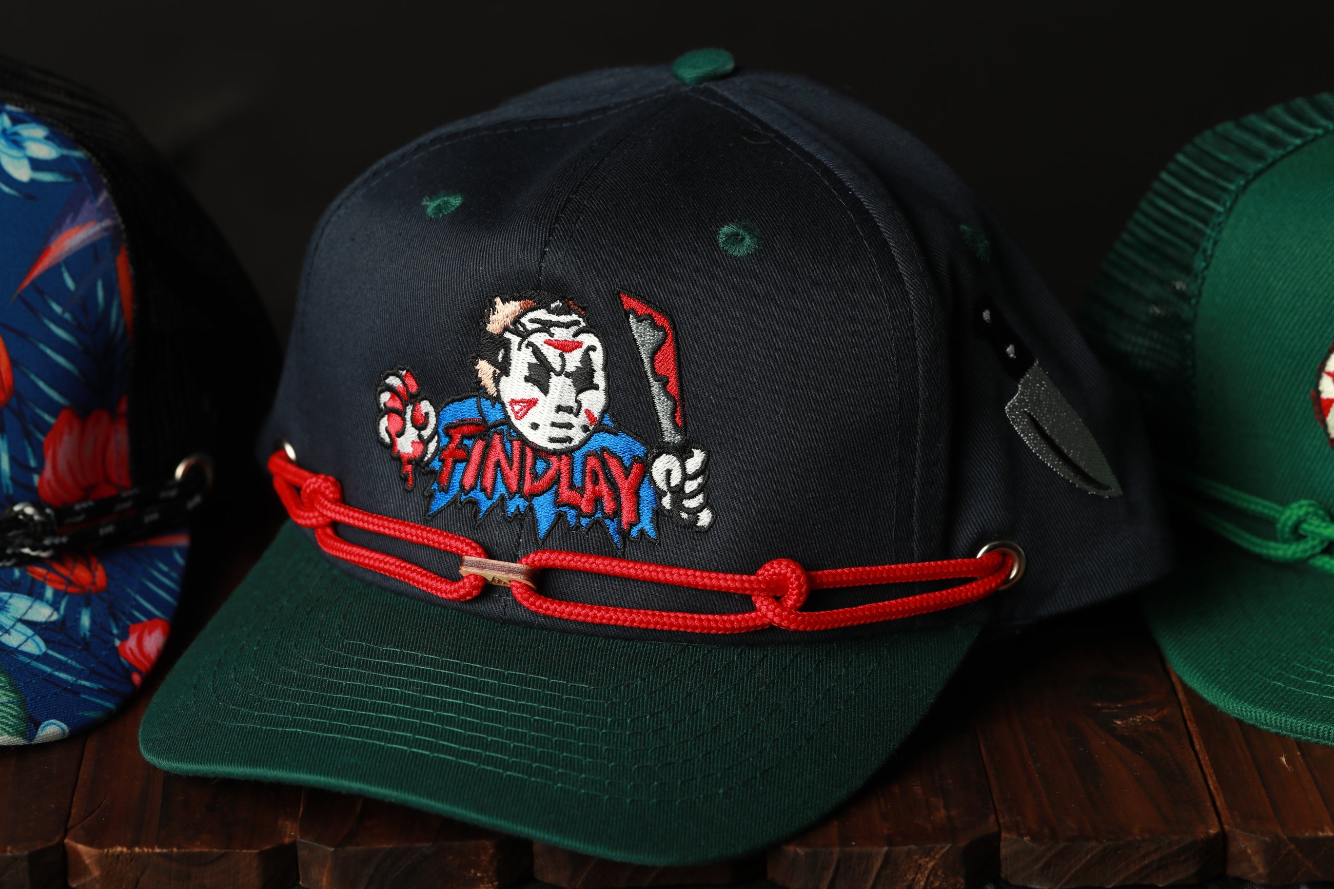 A black baseball cap with a red string and a graphic of a character wearing a mask, along with the text "FINDLAY".