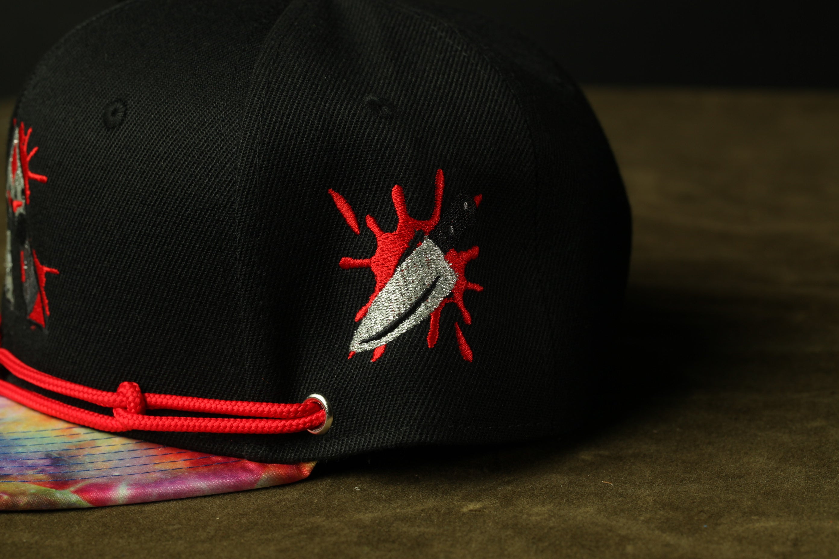 A black baseball cap with a red and white embroidered design on the front, featuring a knife with a red splatter.