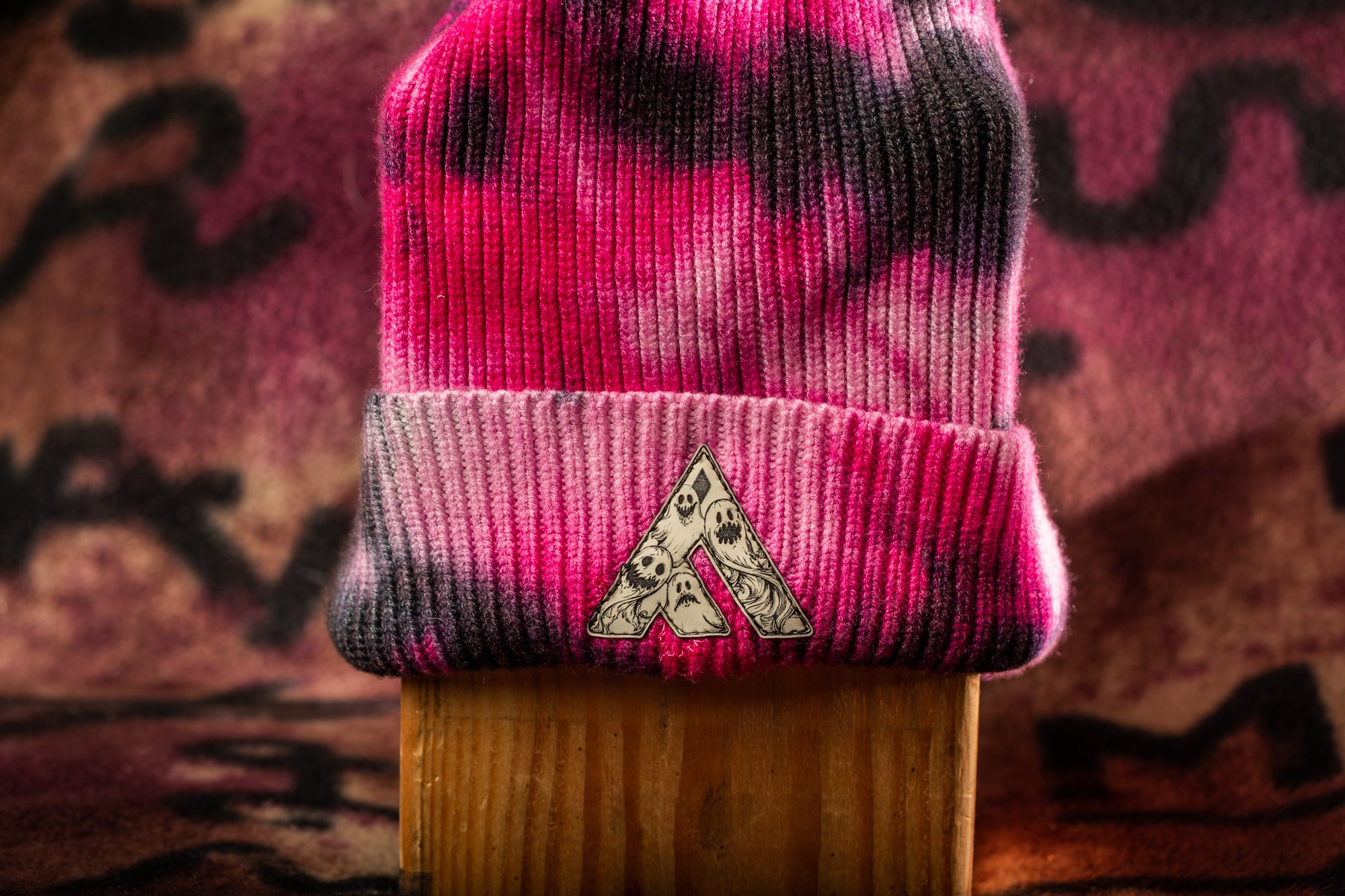 A tie-dye knit beanie with a logo on it, placed on a wooden surface against a colorful background.