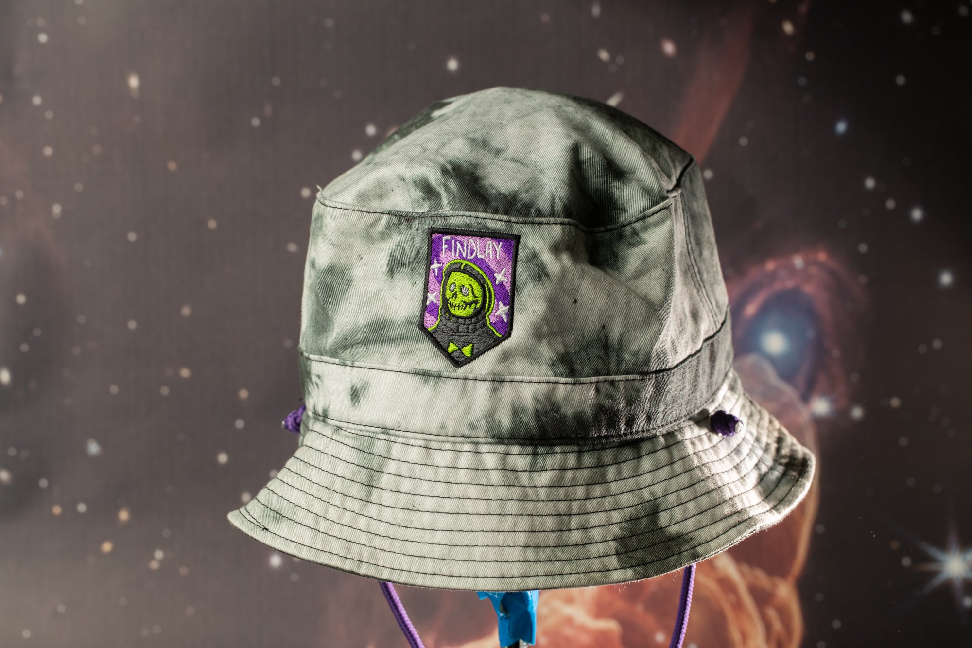 A gray bucket hat with a purple and green patch featuring a skull and crossbones design, set against a backdrop of a starry night sky.