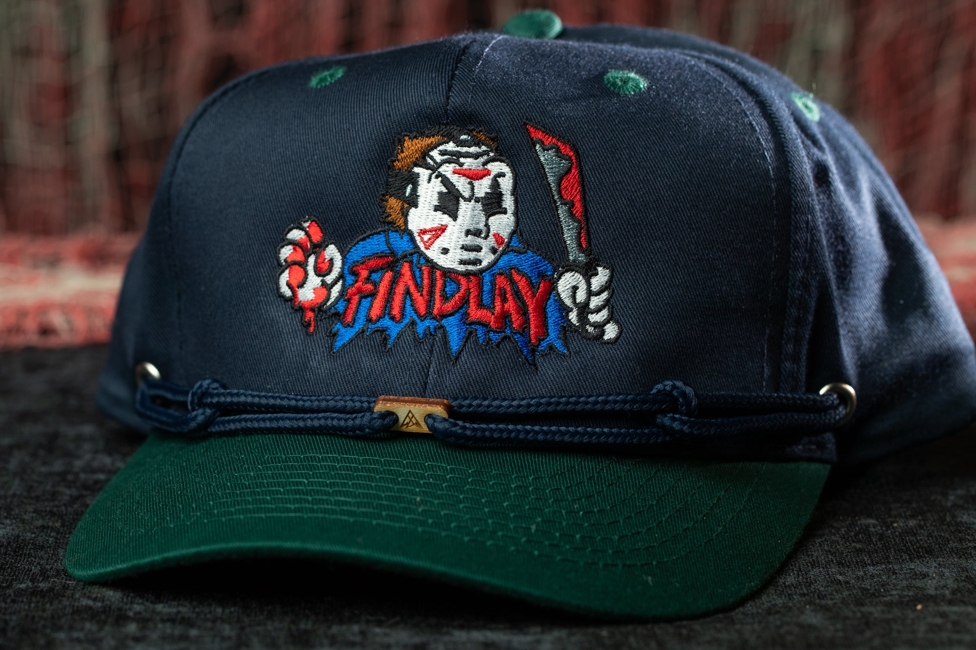 A dark blue baseball cap with a green brim, featuring a cartoon character and the word "FINDLAY" printed on it.
