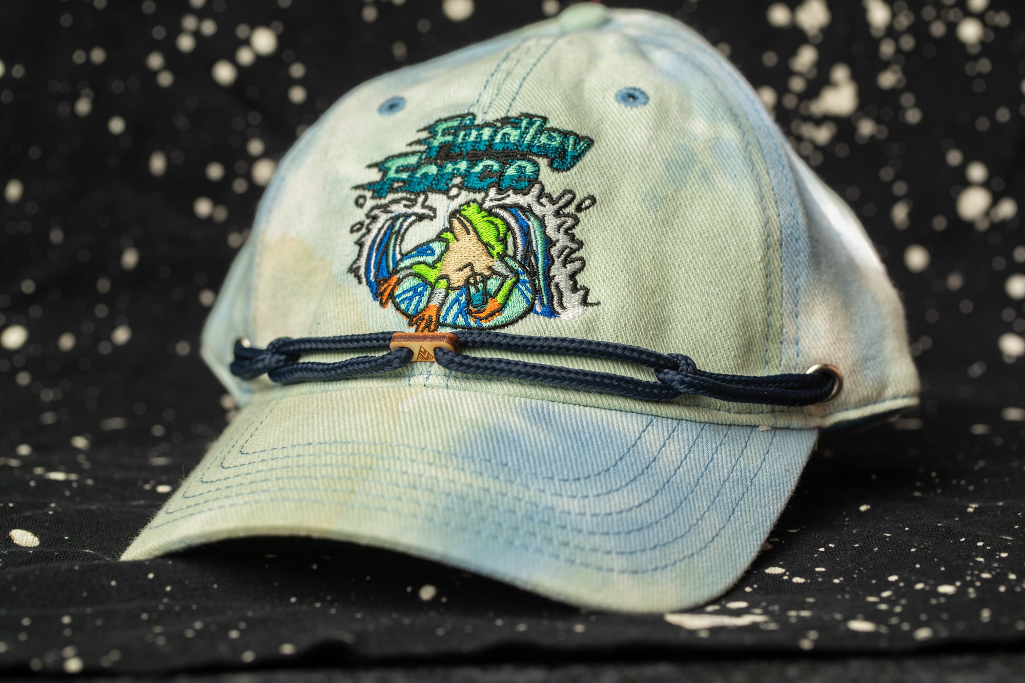 A blue baseball cap with a tie-dye design and the text "Fishing Force" printed on it, sitting on a black background with white speckles.