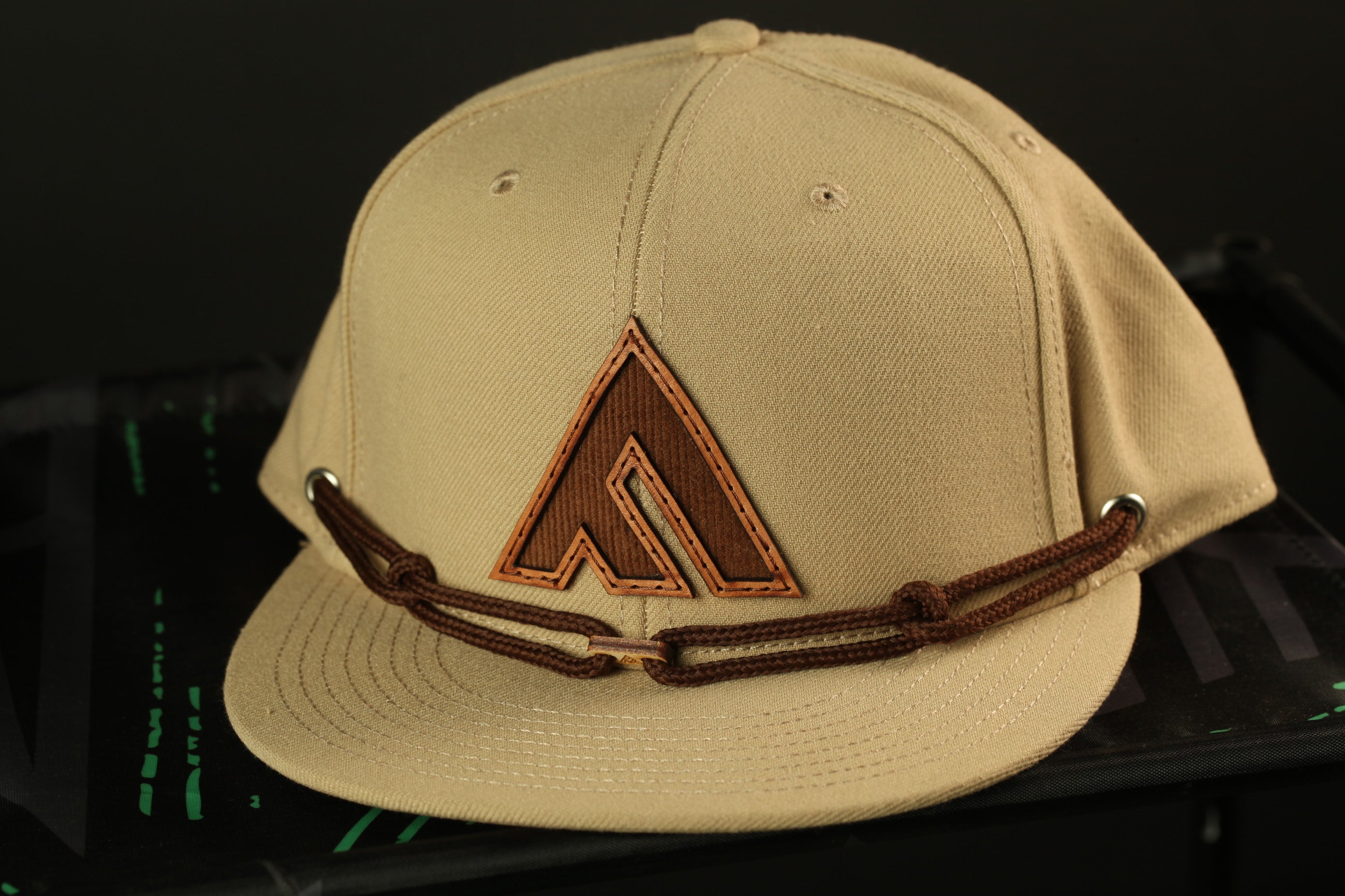 A beige baseball cap with a brown logo on the front, sitting on a black surface.