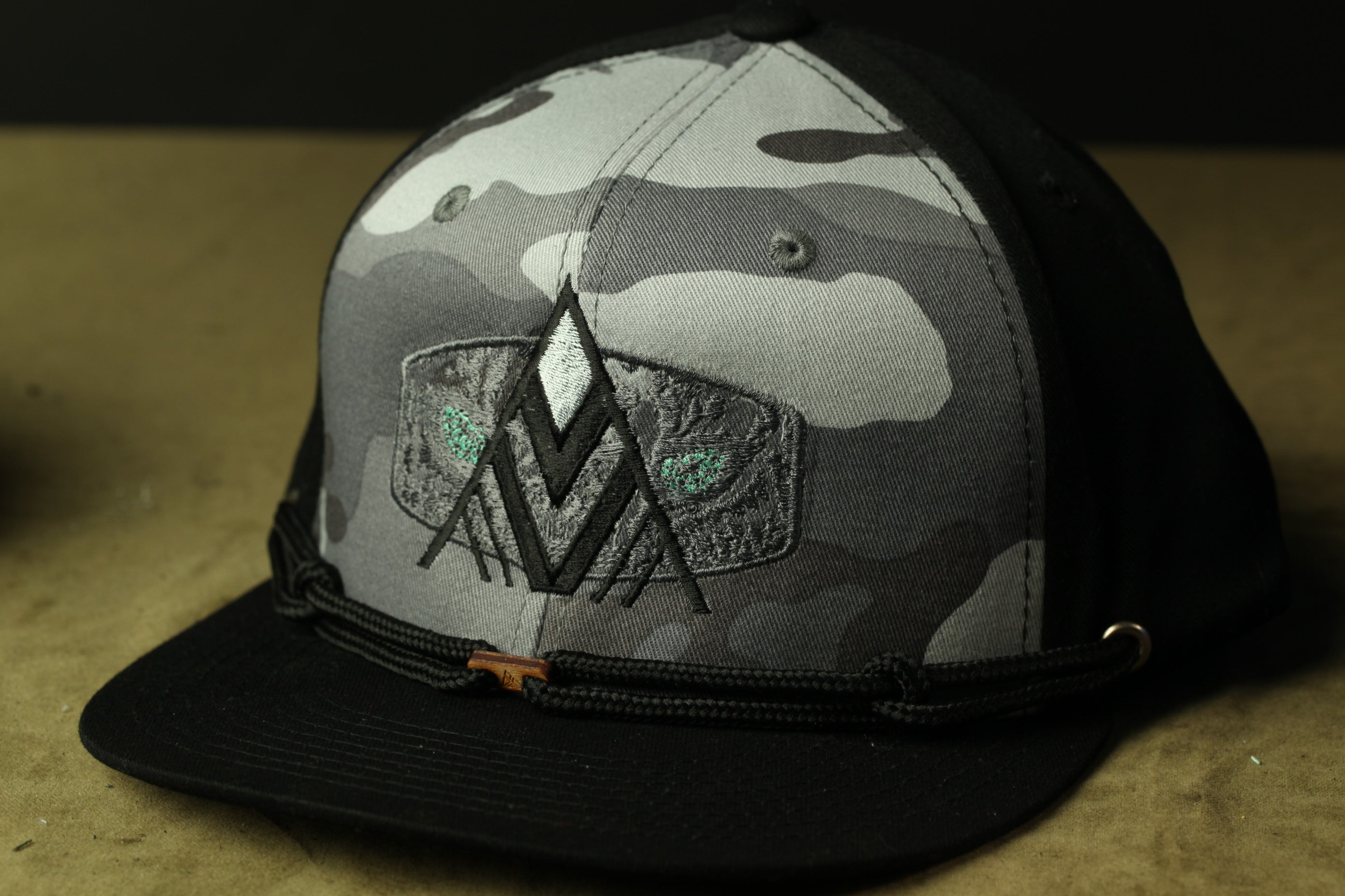 A black and gray camouflage baseball cap with a distinctive logo on the front.