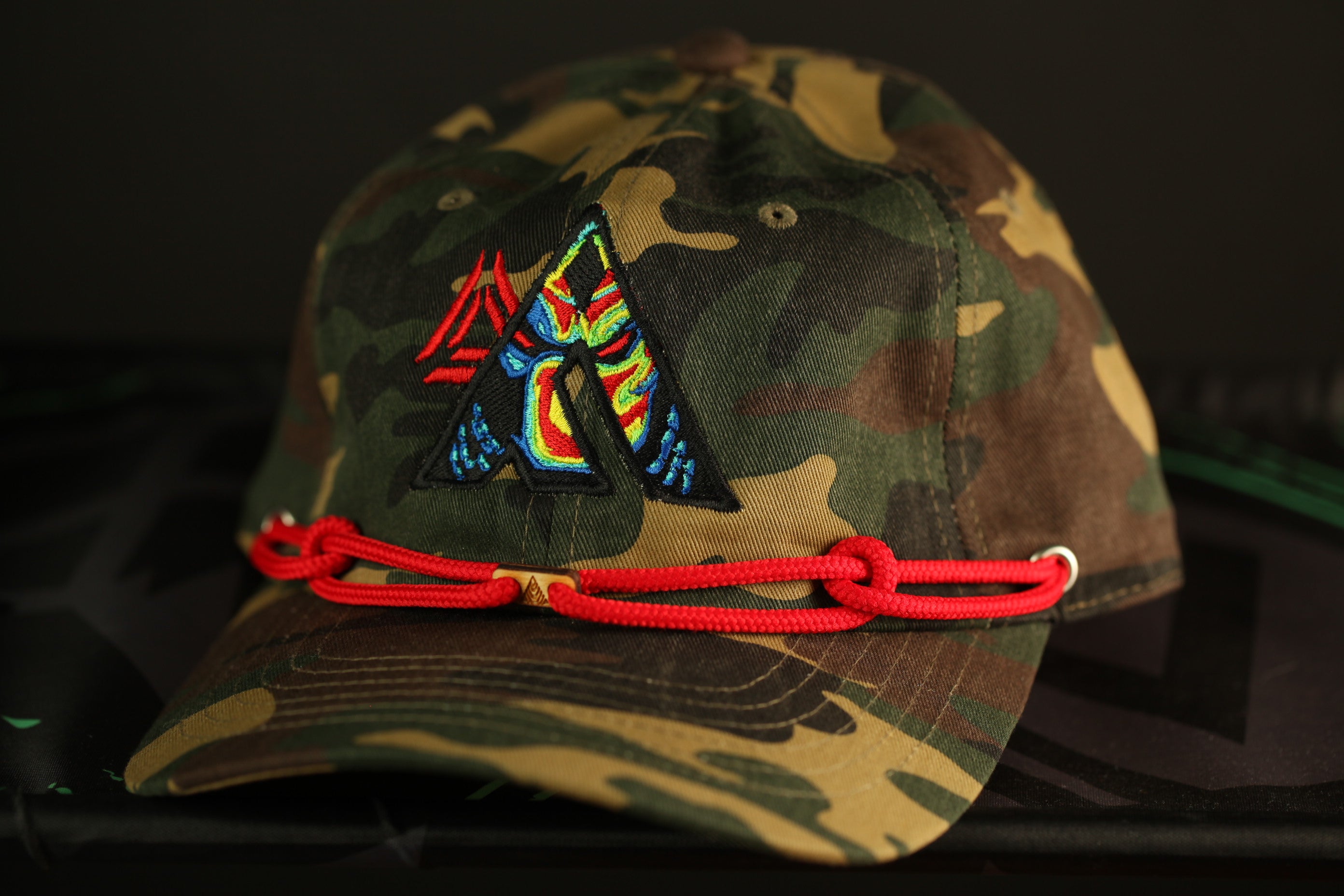 A camouflage baseball cap with a red rope and a colorful triangle-shaped logo on the front.