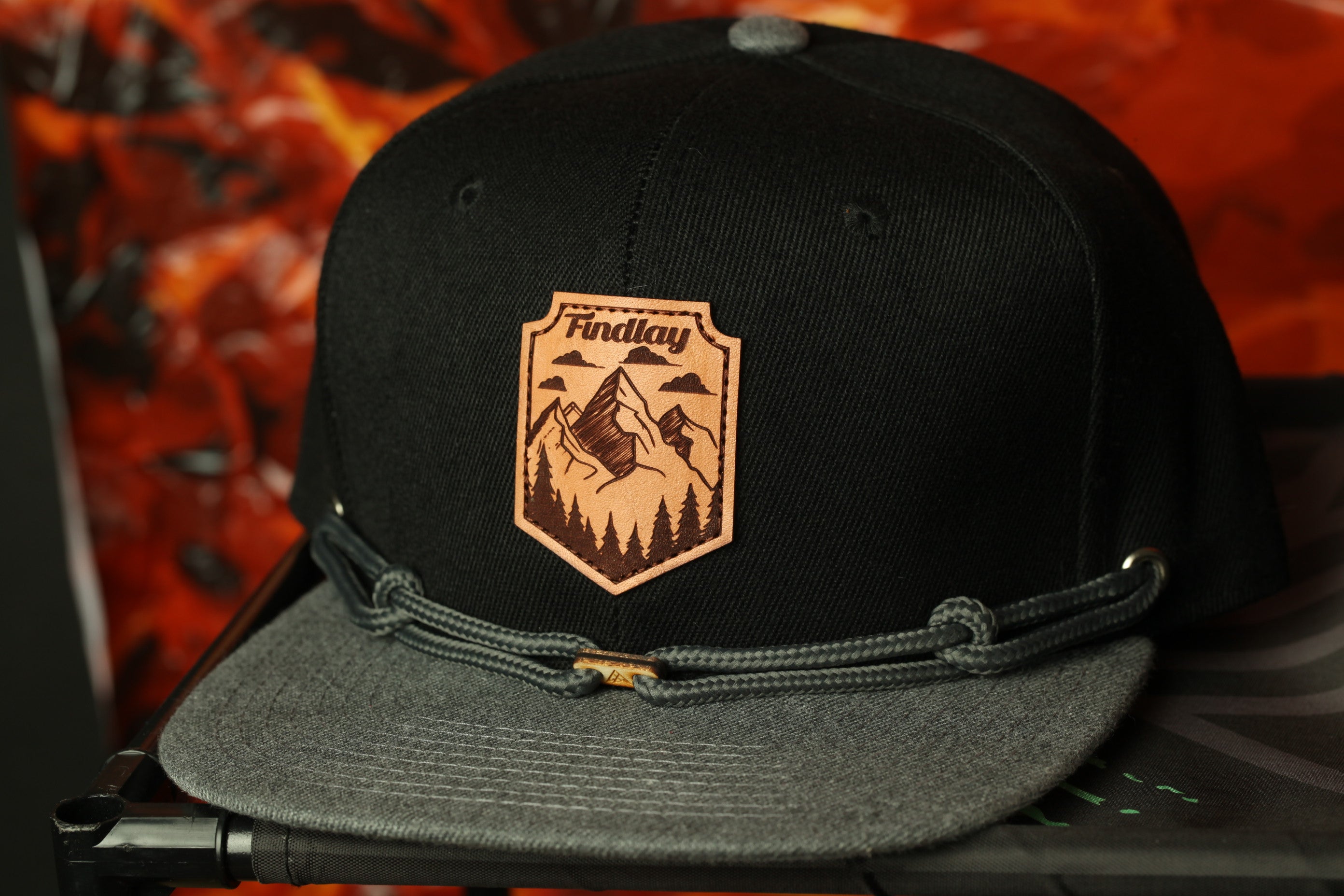 A black baseball cap with a gray brim and a leather patch featuring a mountain landscape.