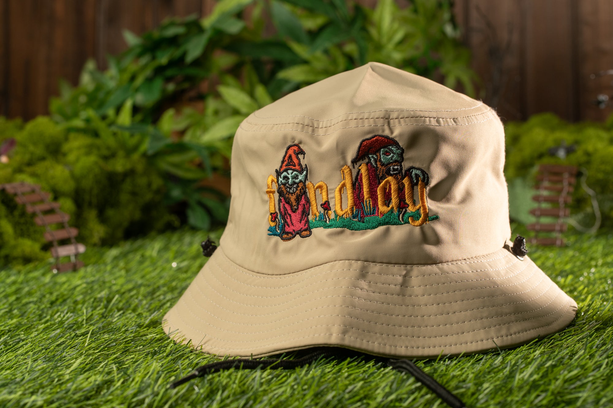 A beige bucket hat with a cartoon character design sits on a grassy surface, with a wooden fence and greenery in the background.