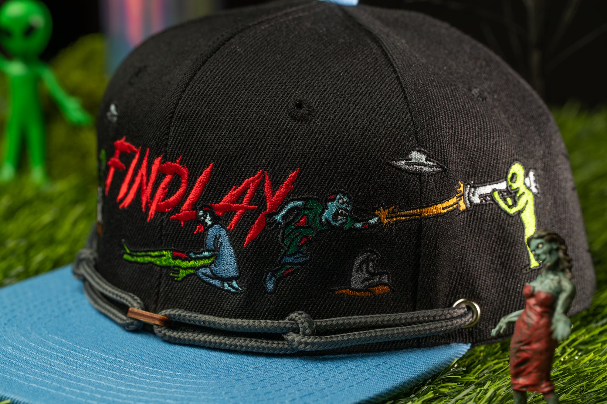 A black baseball cap with a colorful, embroidered design featuring characters and text, placed on a grassy surface with small figurines in the background.