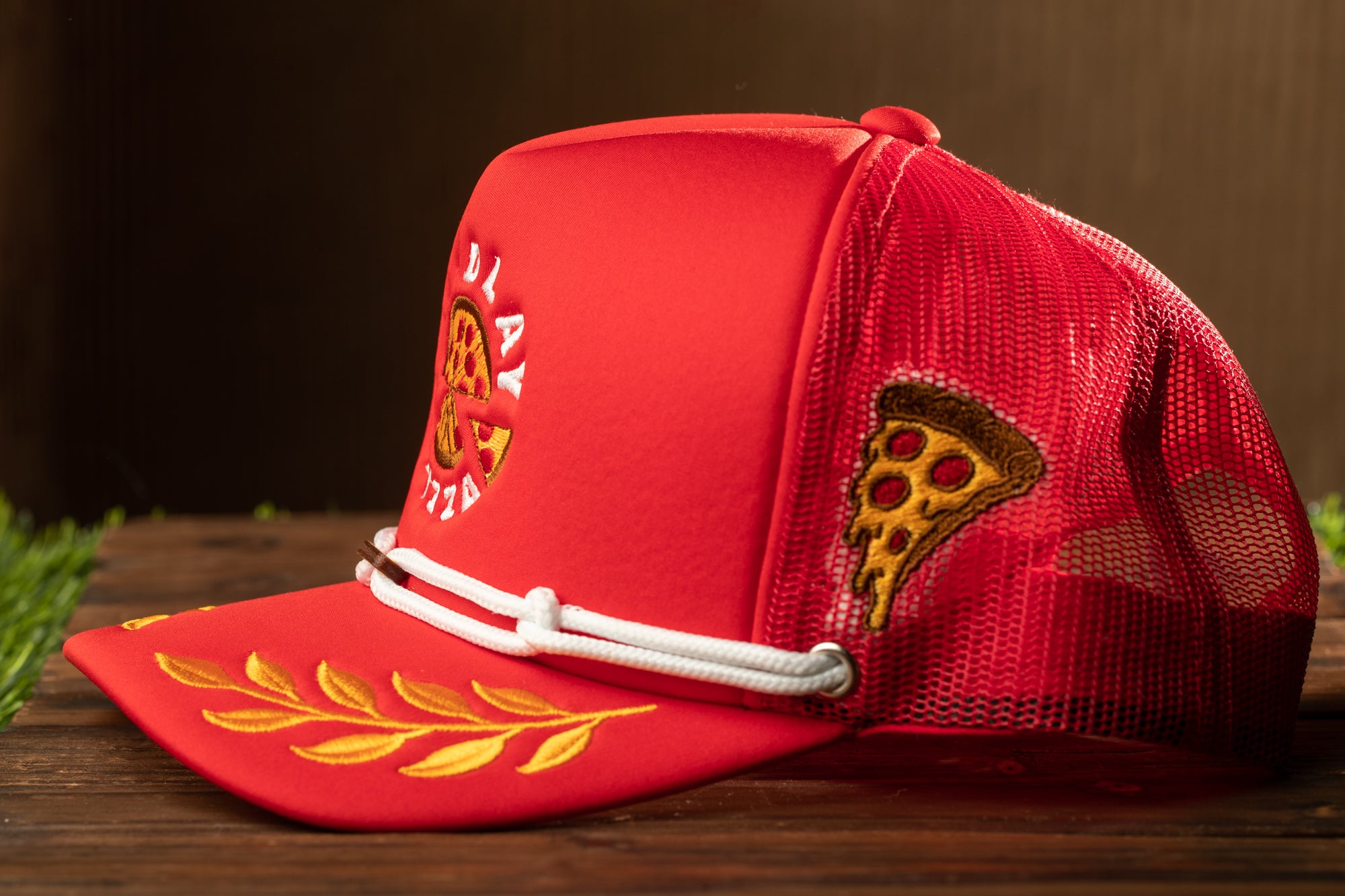 A red baseball cap with a white lace detail and a pizza slice embroidered on the side, sitting on a wooden surface.