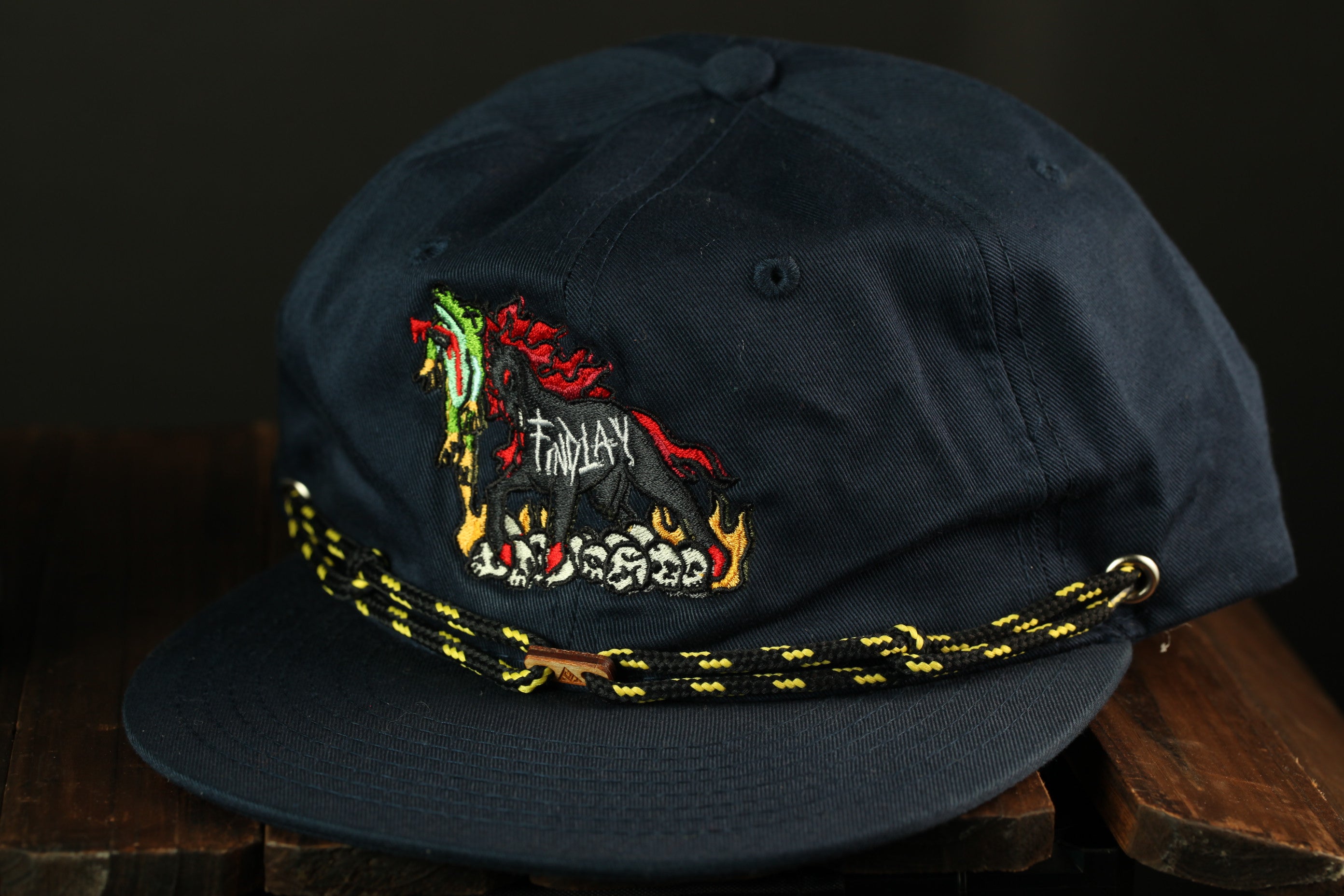 A navy blue baseball cap with a colorful embroidered design on the front, featuring a stylized bird or creature.