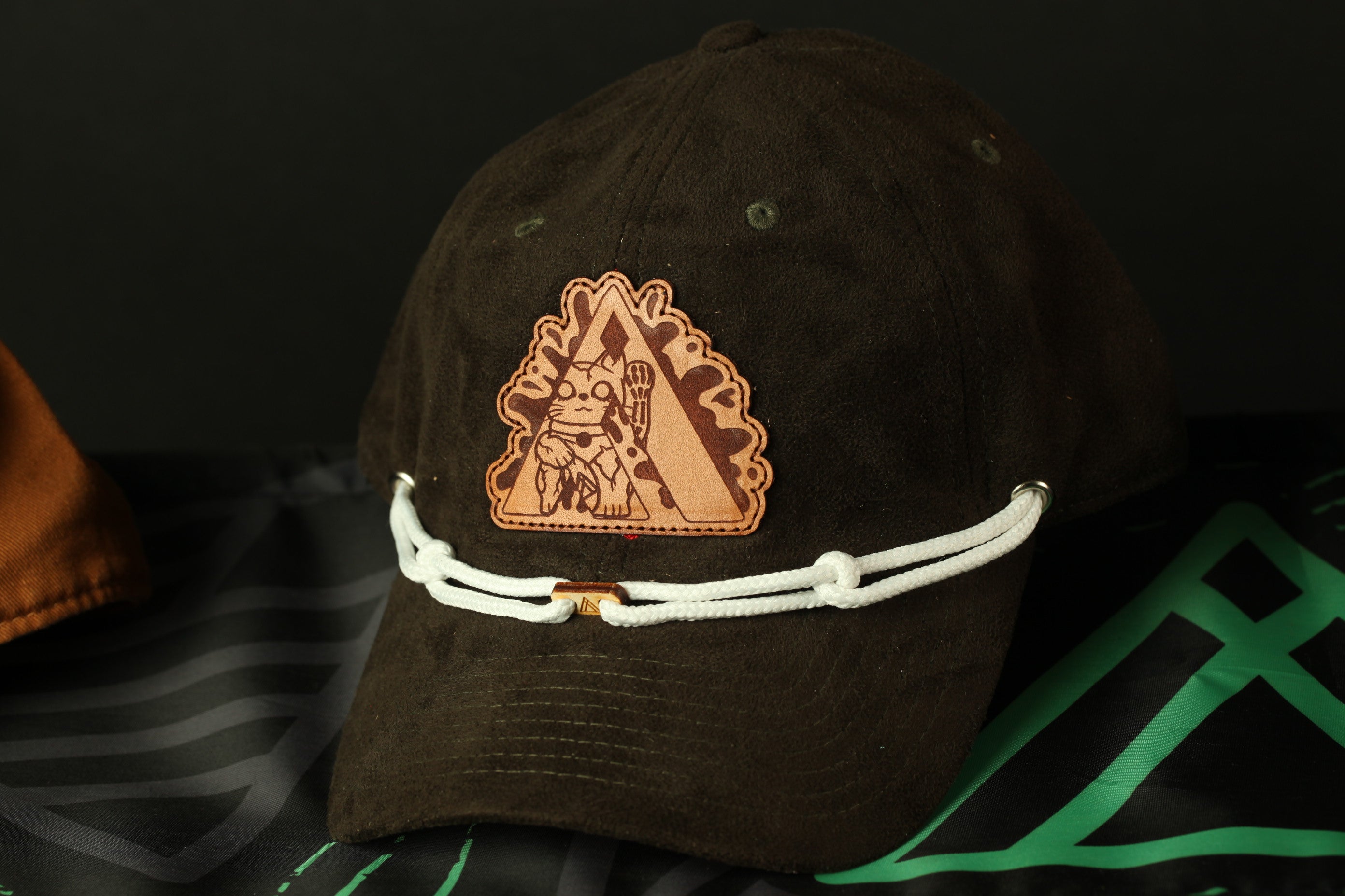 A brown baseball cap with a white string and a leather patch featuring a cartoon character is displayed against a black background.