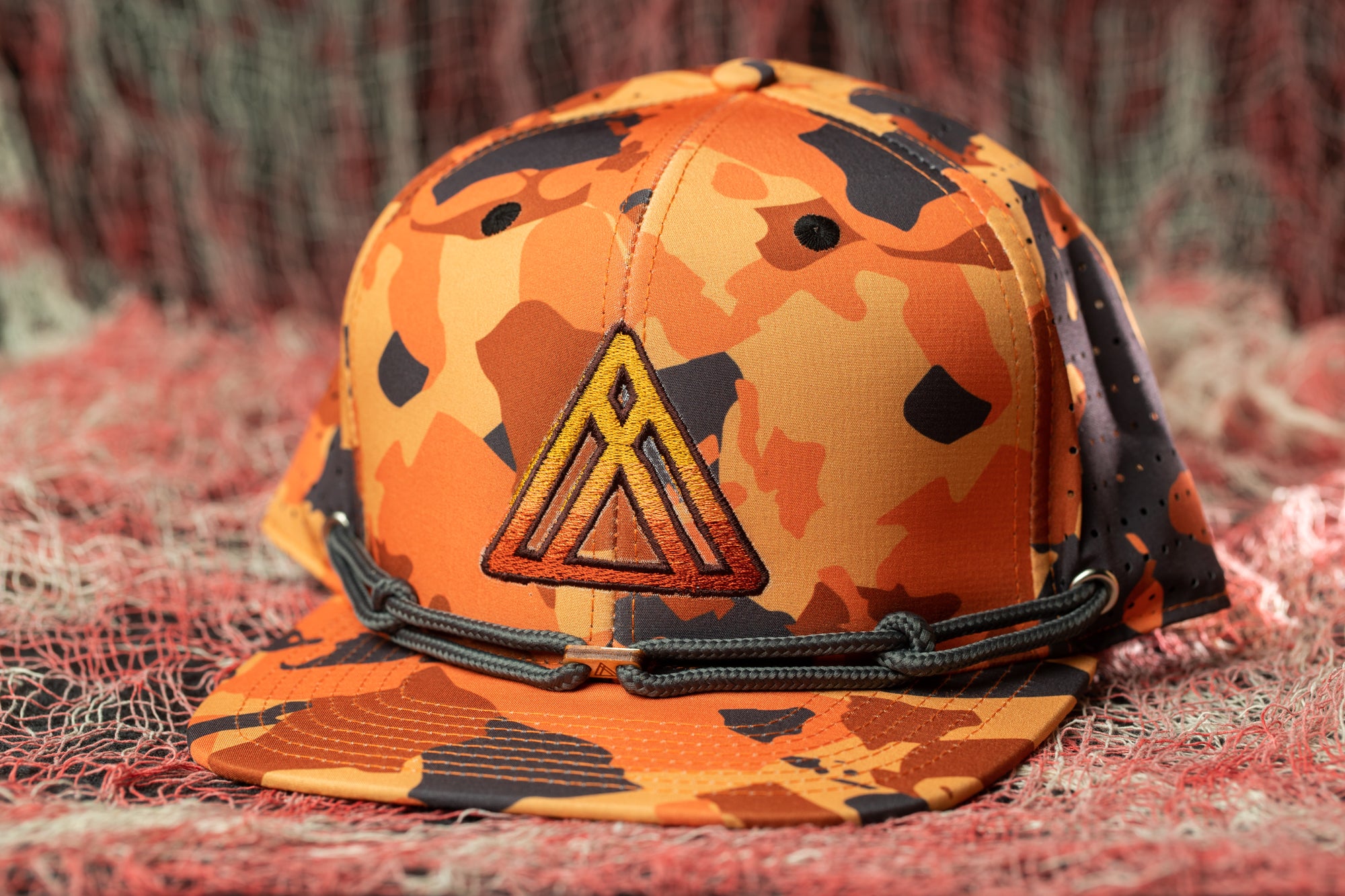 A camouflage baseball cap with a triangular logo on the front, resting on a red and white textured surface.