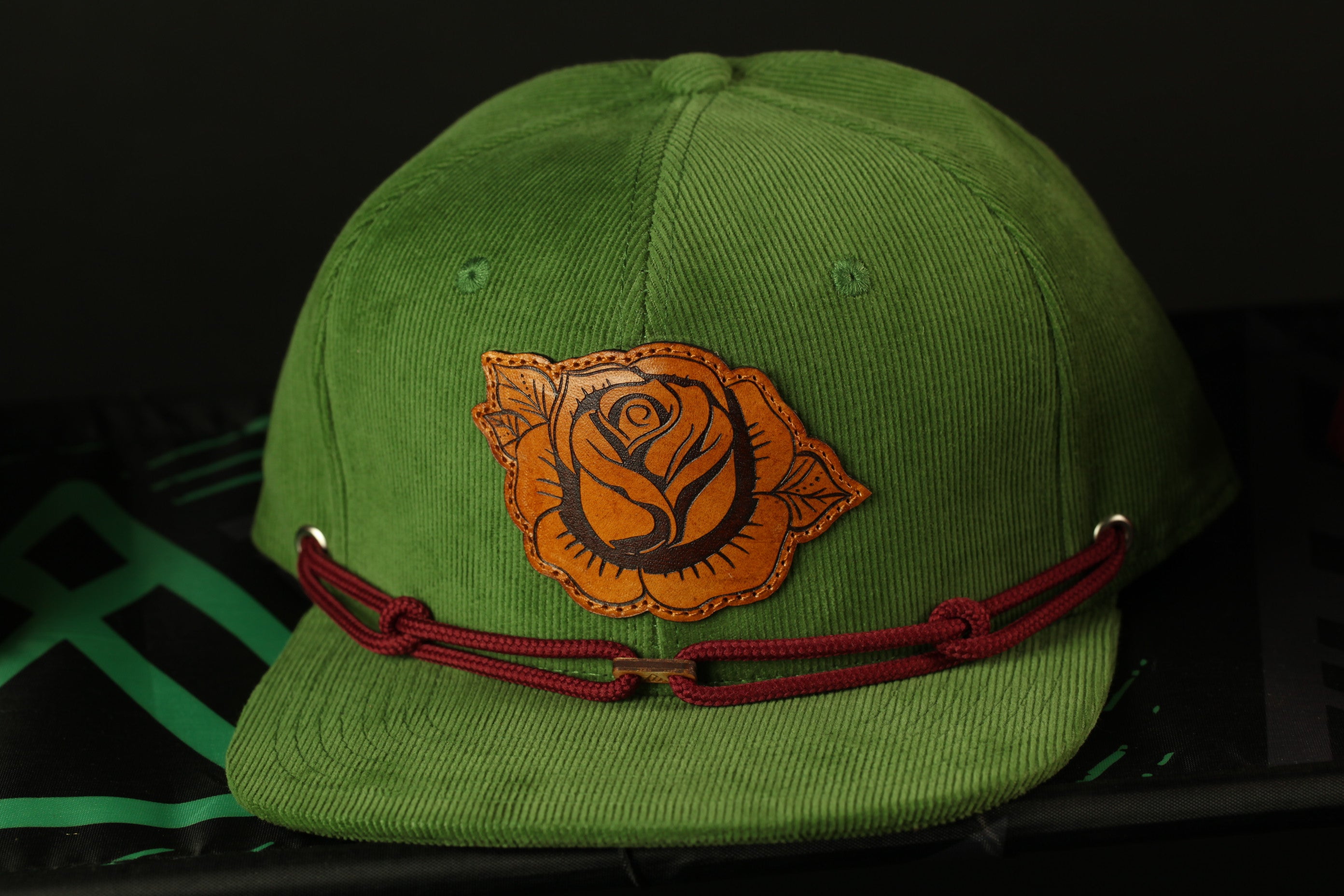 A green baseball cap with a rose embroidered on the front and a red lace strap.