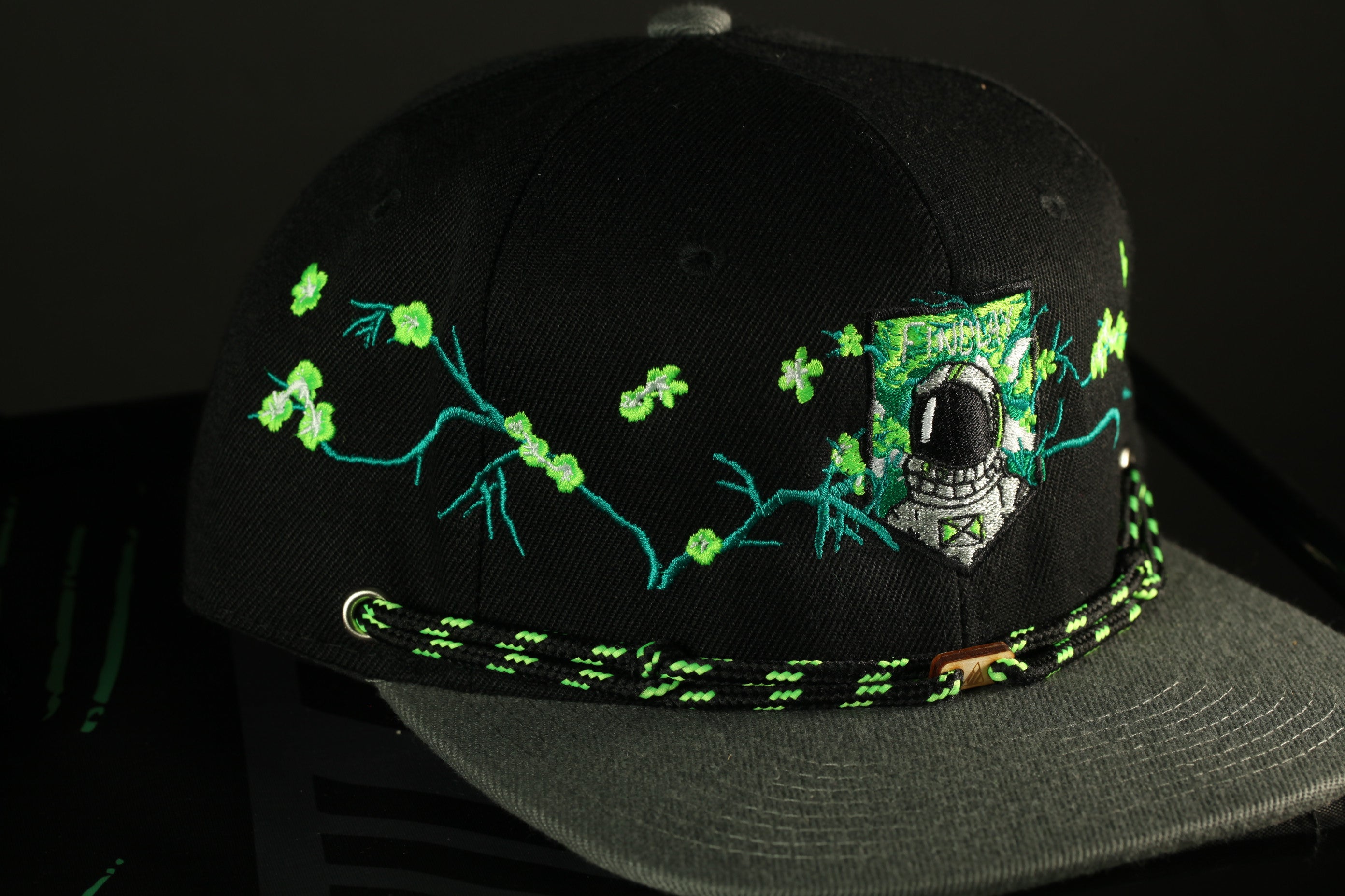 A black baseball cap with a green and black braided strap, featuring a patch with a skull and crossbones design on the front.