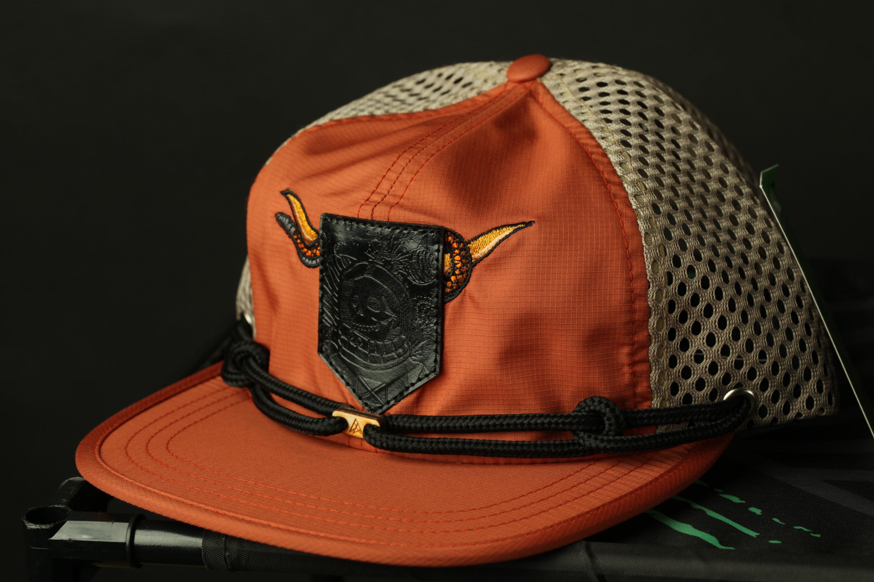 An orange baseball cap with a mesh panel and a black leather patch on the front, featuring a dragon design.