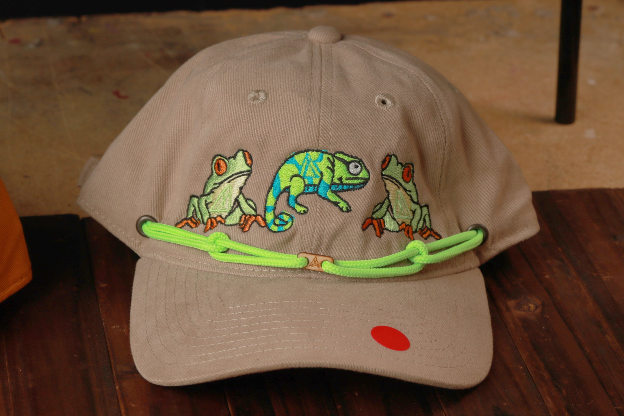 A beige baseball cap with a green string and a red dot, featuring a design of three green chameleons.