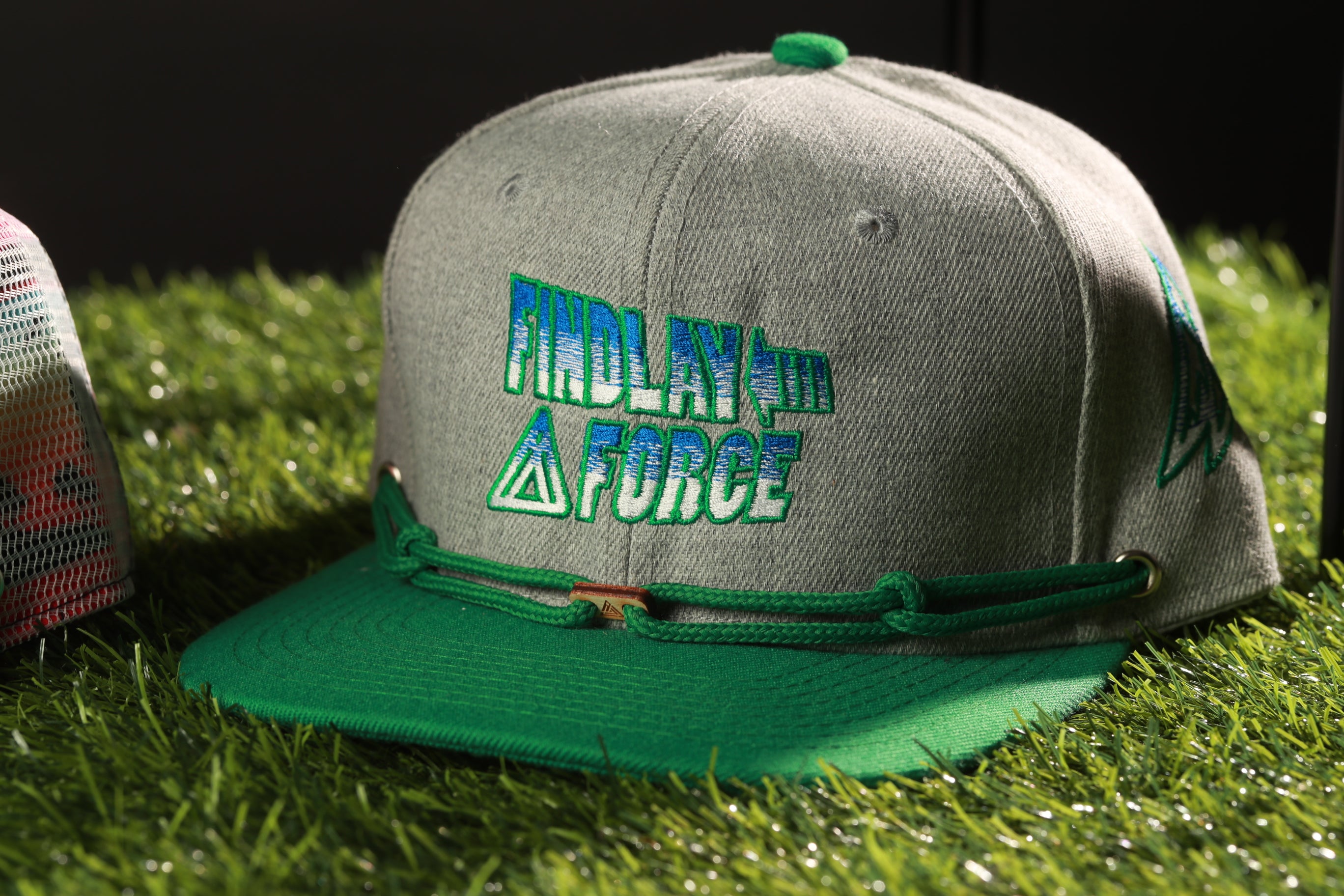 A gray baseball cap with a green brim and a logo on it, placed on a grassy surface.