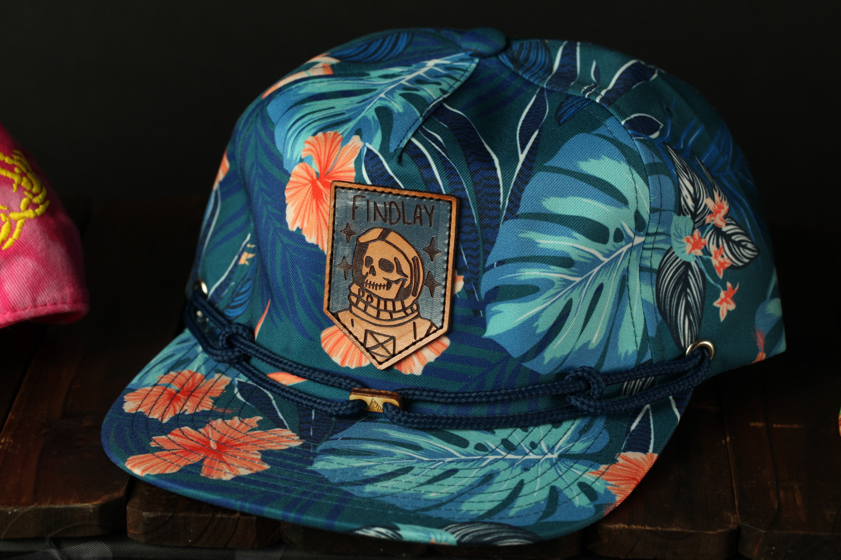A colorful tropical-themed baseball cap with a skull patch and a blue band, displayed on a wooden surface.